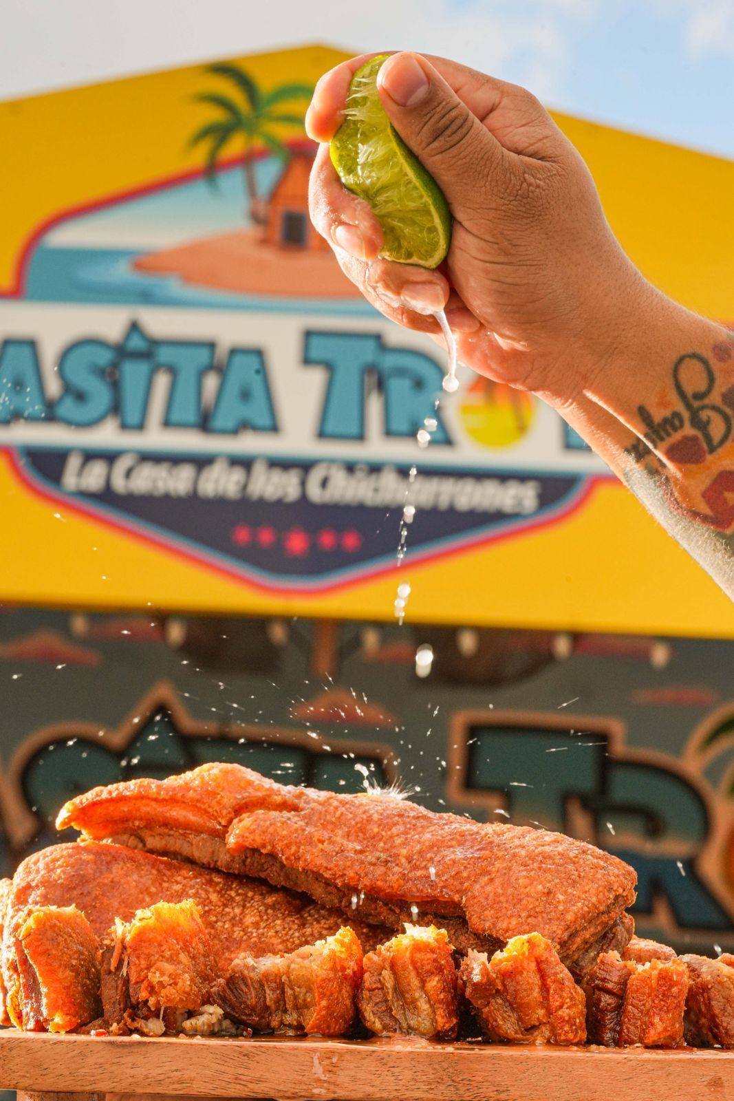 A hand squeezes lime juice over crispy chicharrón. A brightly colored food stand with 