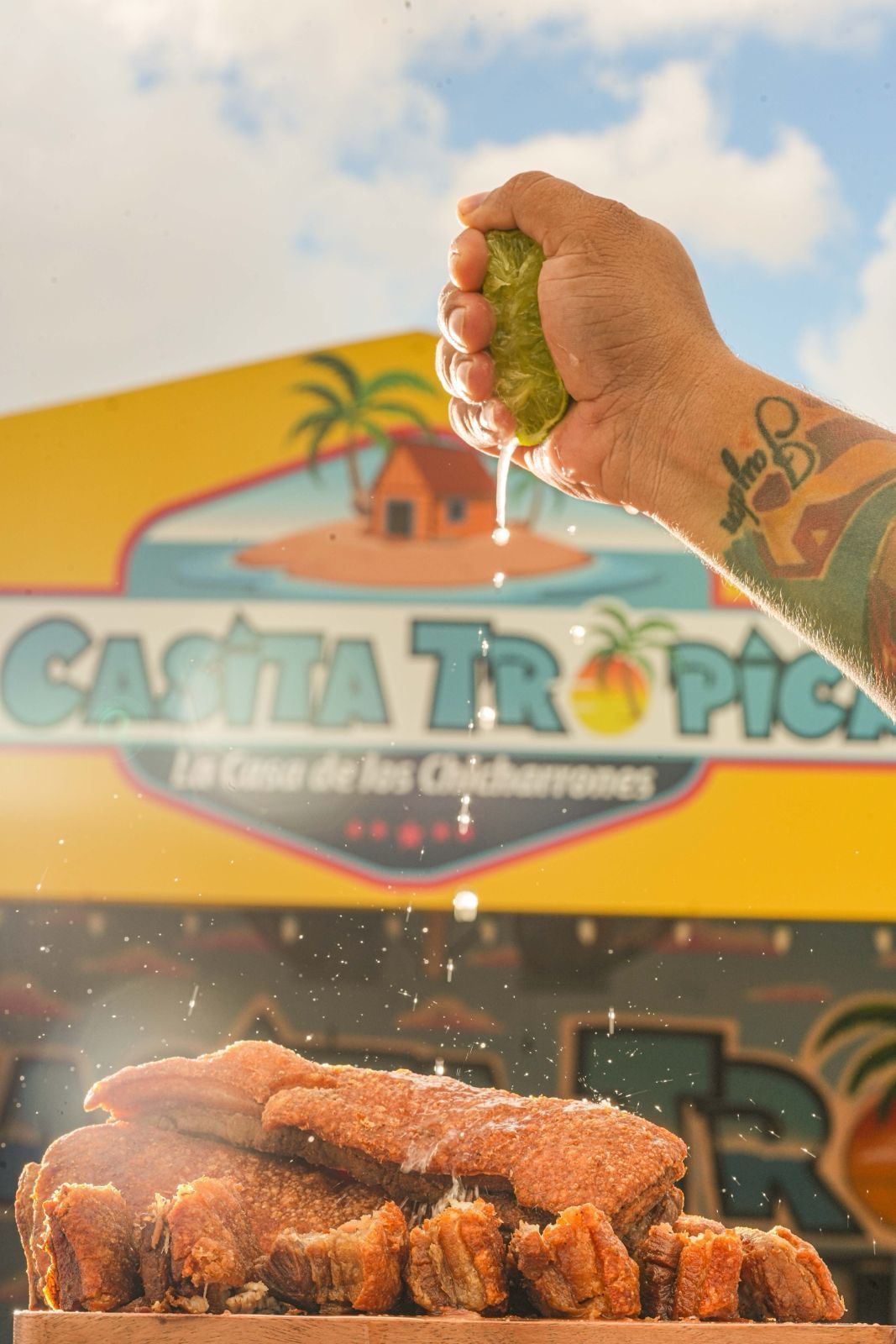 A hand squeezes lime juice over fried pork rinds in front of a tropical-themed food stand, 