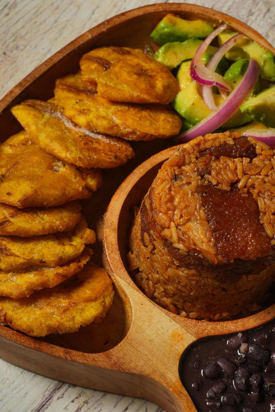 Wooden plate with tostones, avocado and red onion salad, rice, and black beans.
