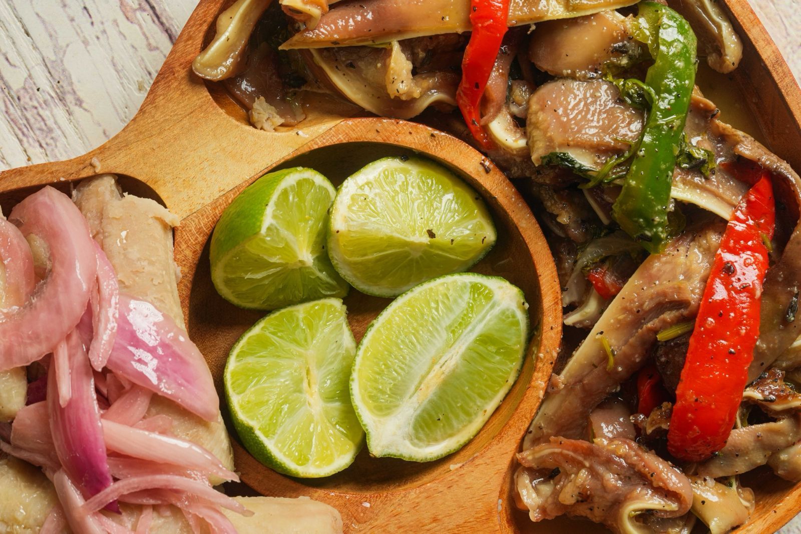 Wooden plate with lime wedges, pickled onions, and cooked meat with peppers.