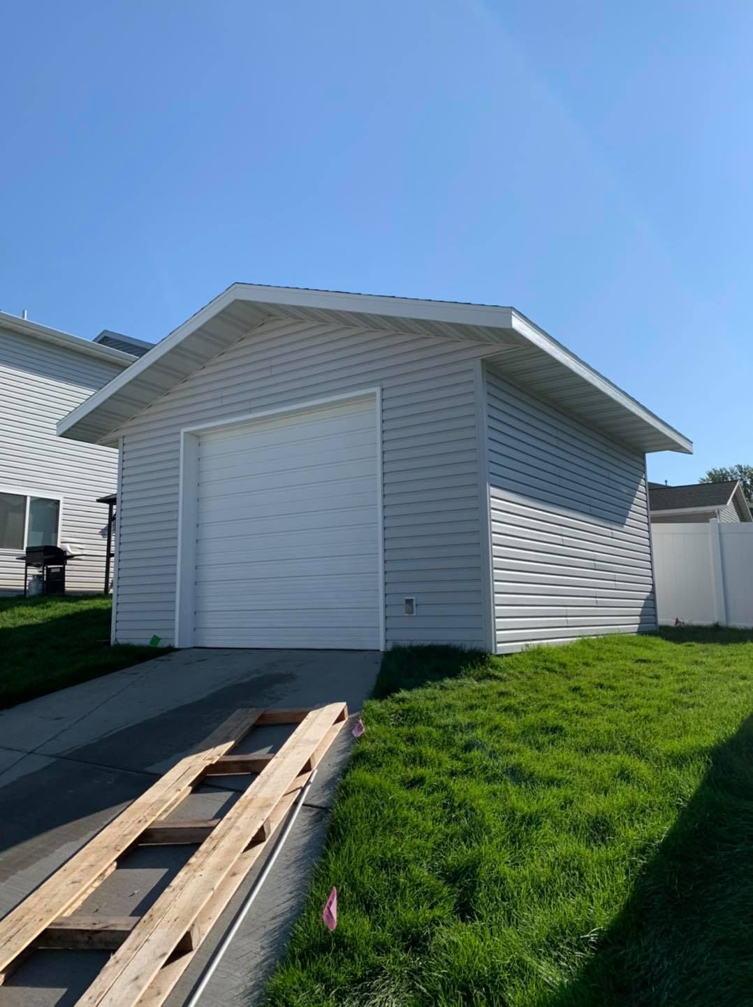 Garage with a white door and gray siding, on a grassy lot. Wooden planks in foreground.