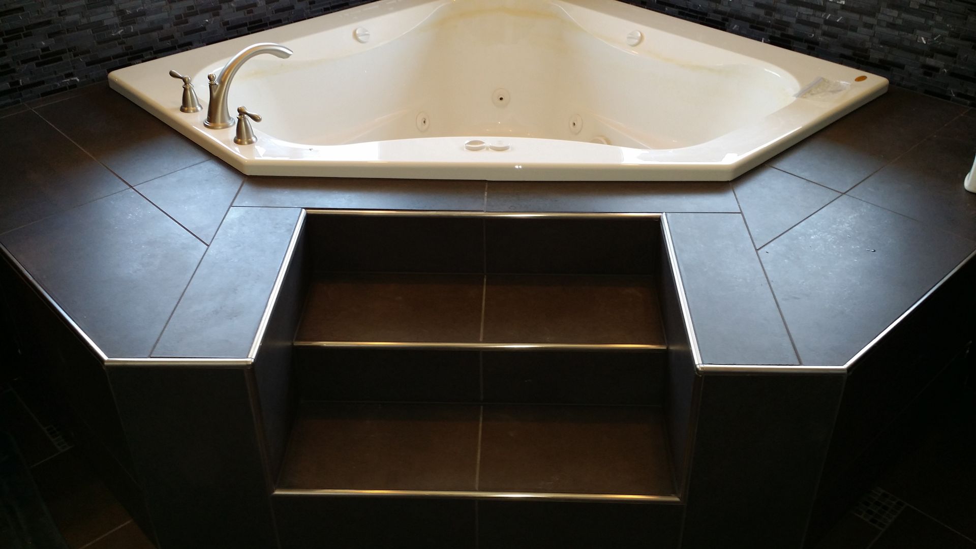 Corner jacuzzi tub with tiled surround and built-in shelves. Dark tile, stainless trim.