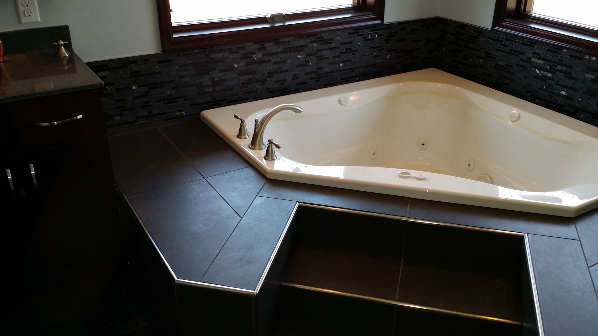 A white corner jacuzzi tub in a dark bathroom. Dark floor and tile wall, window above.