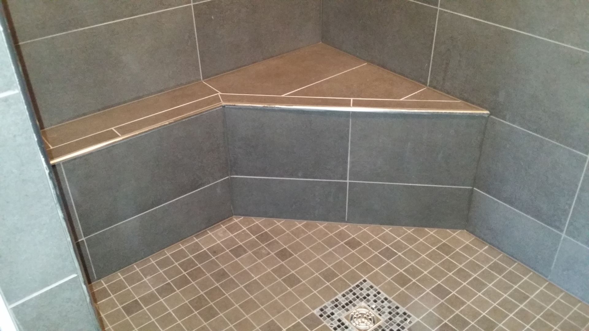Corner shower with gray tile walls and seat. Floor is small brown and tan tiles.