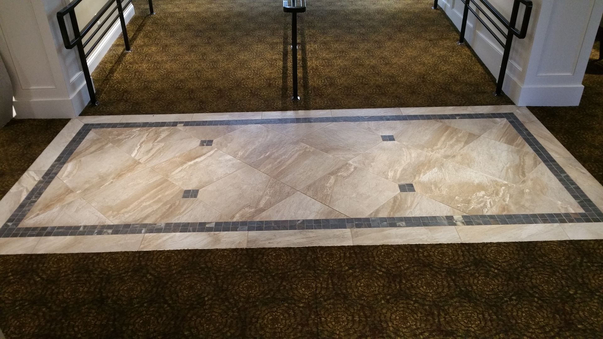 Tiled floor insert with a dark border on a brown carpeted floor, in front of a doorway.