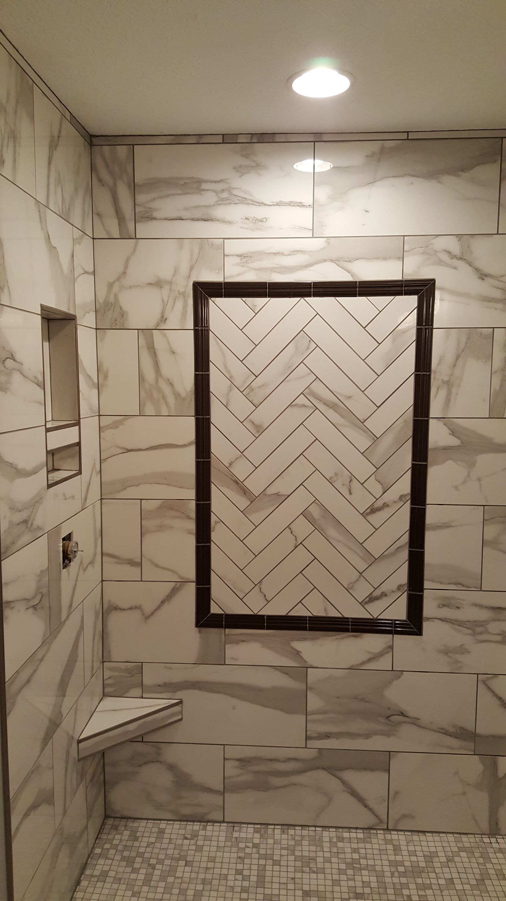 Shower with marble-patterned tiles and herringbone design, featuring a recessed shelf and corner seat.