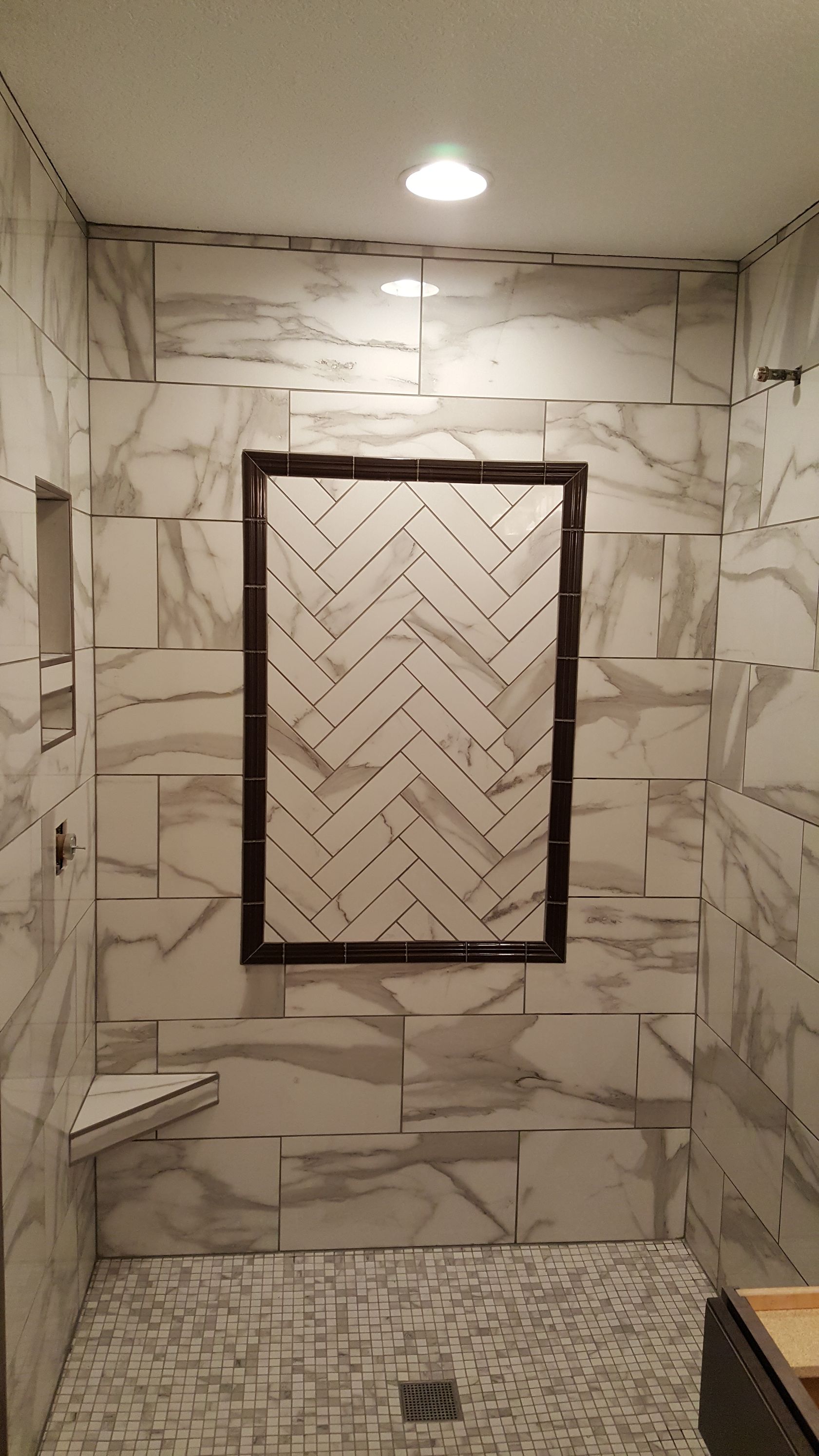 Shower stall with marble-look tiles and a herringbone-patterned inset, featuring a corner seat and a recessed niche.