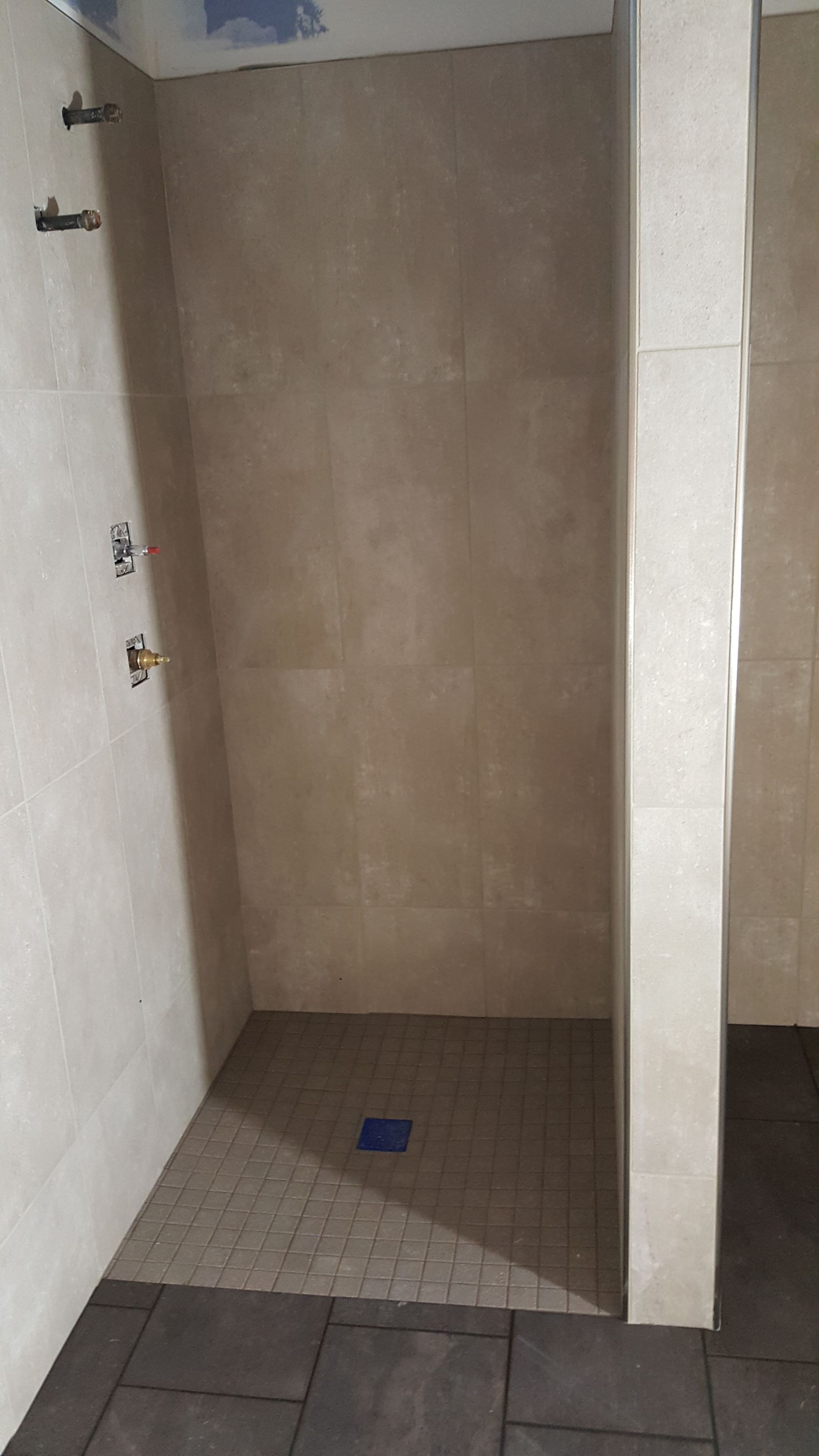 Empty shower stall with light beige tiled walls and dark floor tiles. Shower fixtures are visible on the left wall.