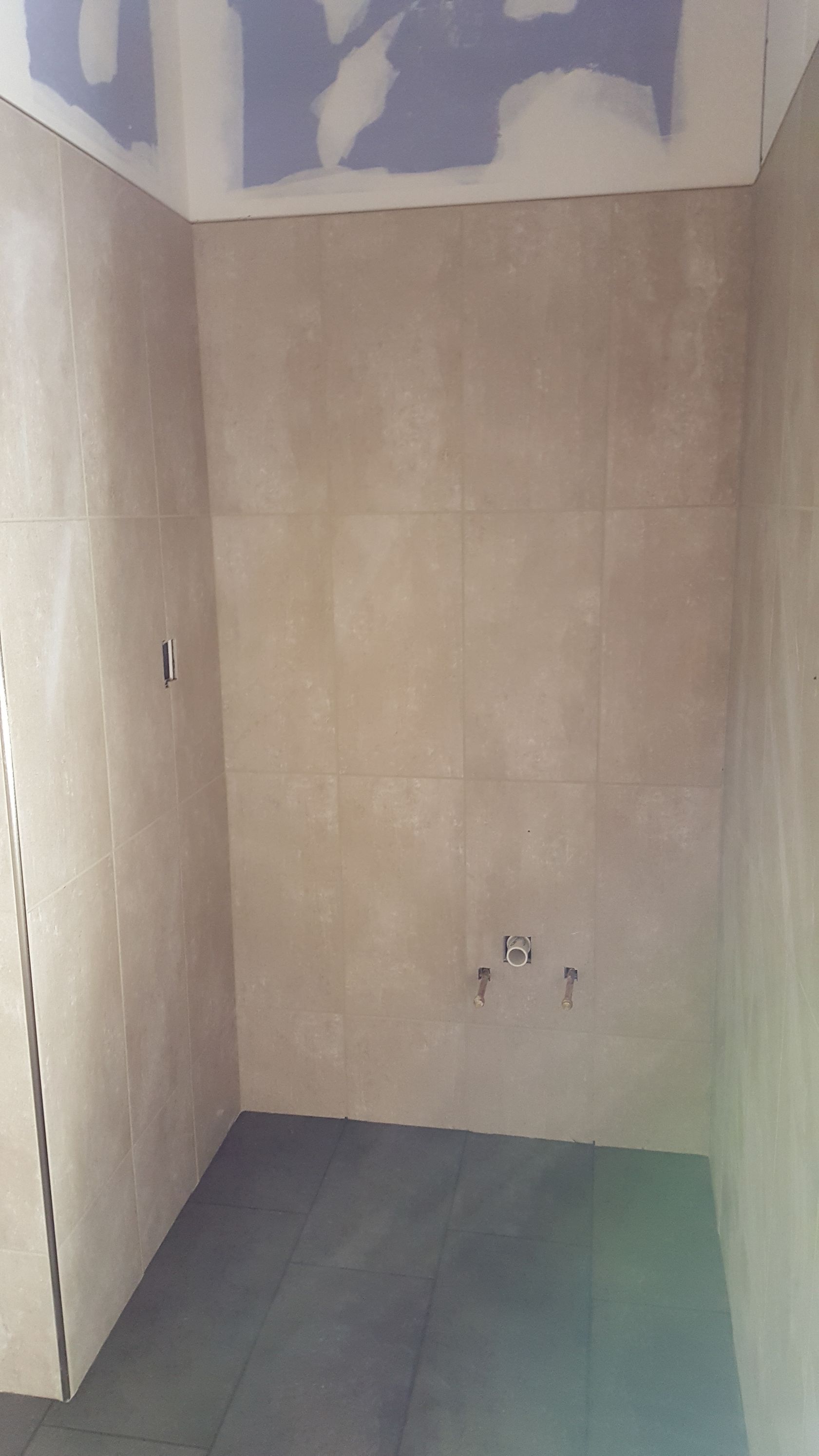 Tiled shower stall in a room under construction; beige walls, dark floor, exposed plumbing.