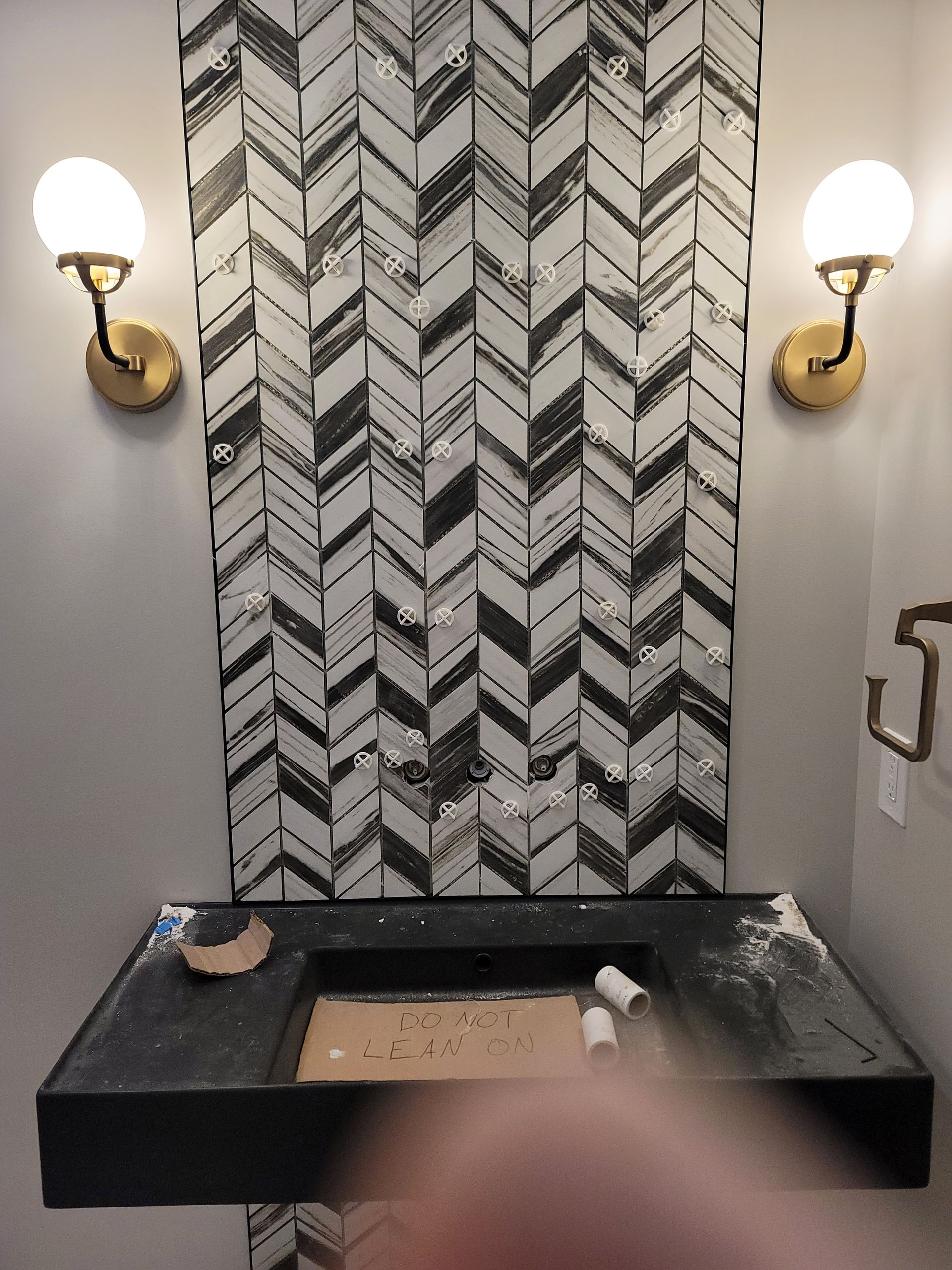 Powder room with herringbone tile backsplash, black counter, gold sconces, and a partially visible countertop.