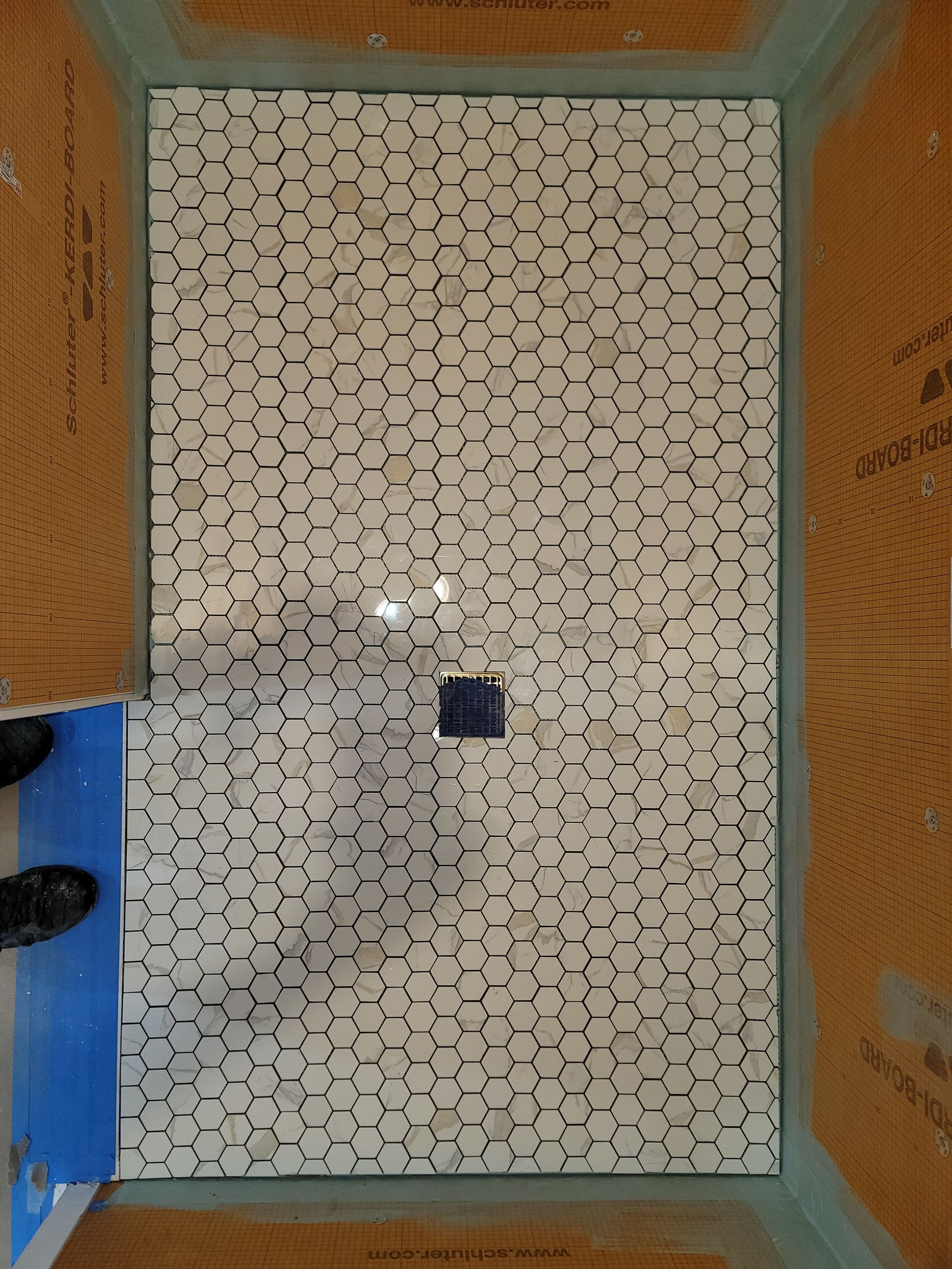 Shower floor with white and gray hexagon tile, drain in center, and surrounding waterproof membrane.