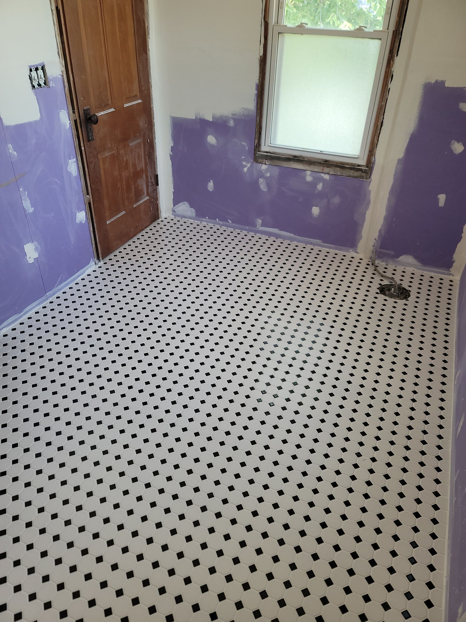 Bathroom renovation in progress; white tile floor with black dots; purple drywall; wooden door and window.