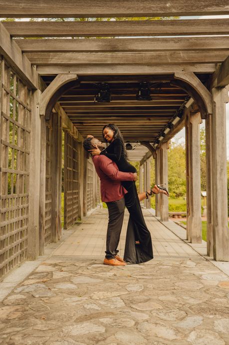 A person in a maroon jacket lifts another person in a long black dress inside a rustic wooden garden pergola.