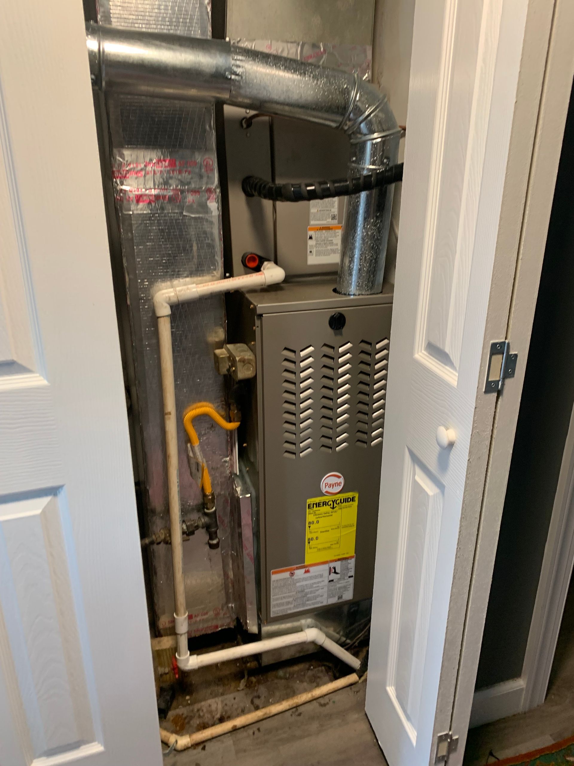 Furnace in a closet, metal and silver with white PVC pipes, yellow gas line.