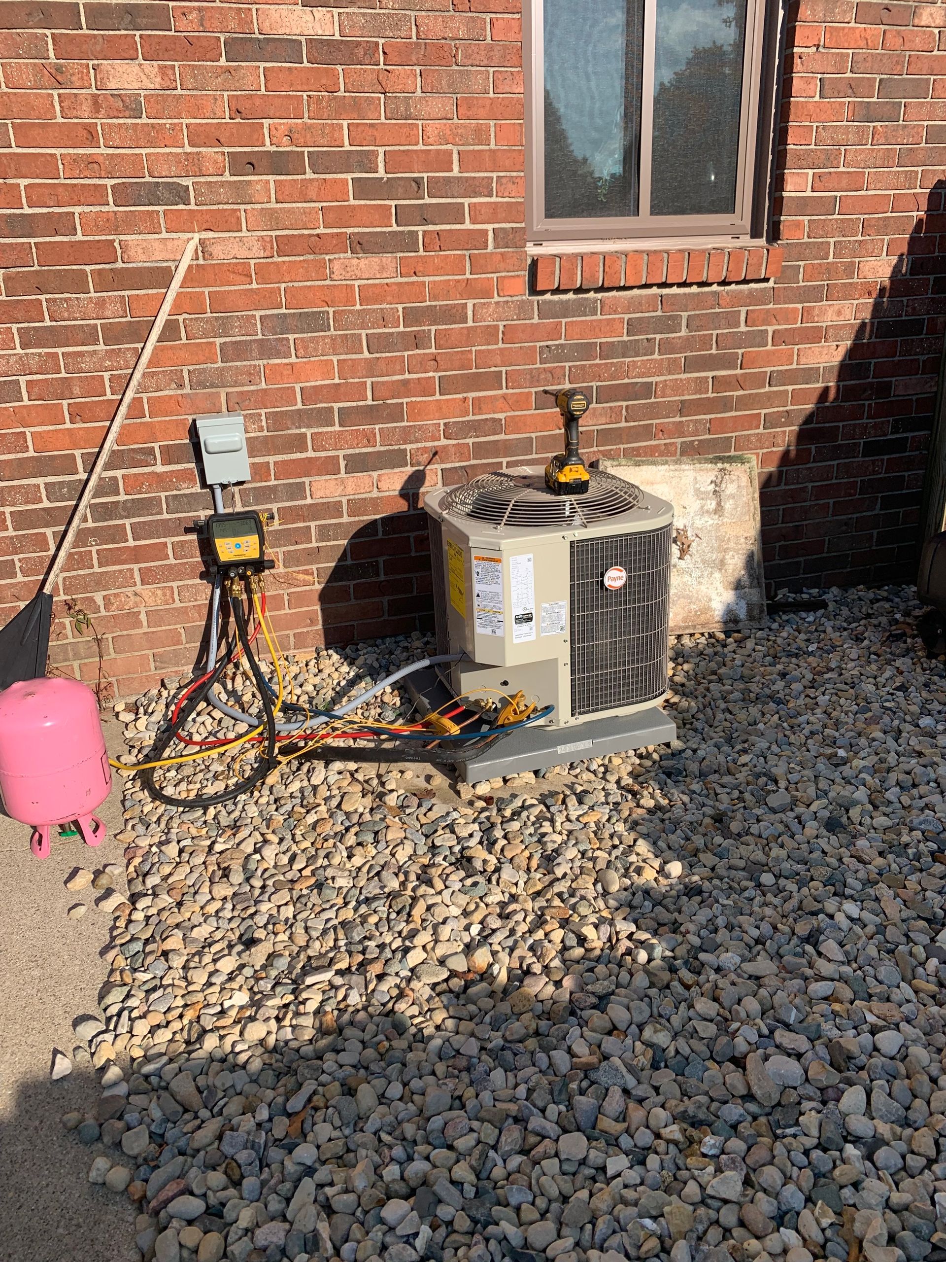 HVAC unit being serviced against a brick wall, tools and gauges connected. Gravel and pink tank present.