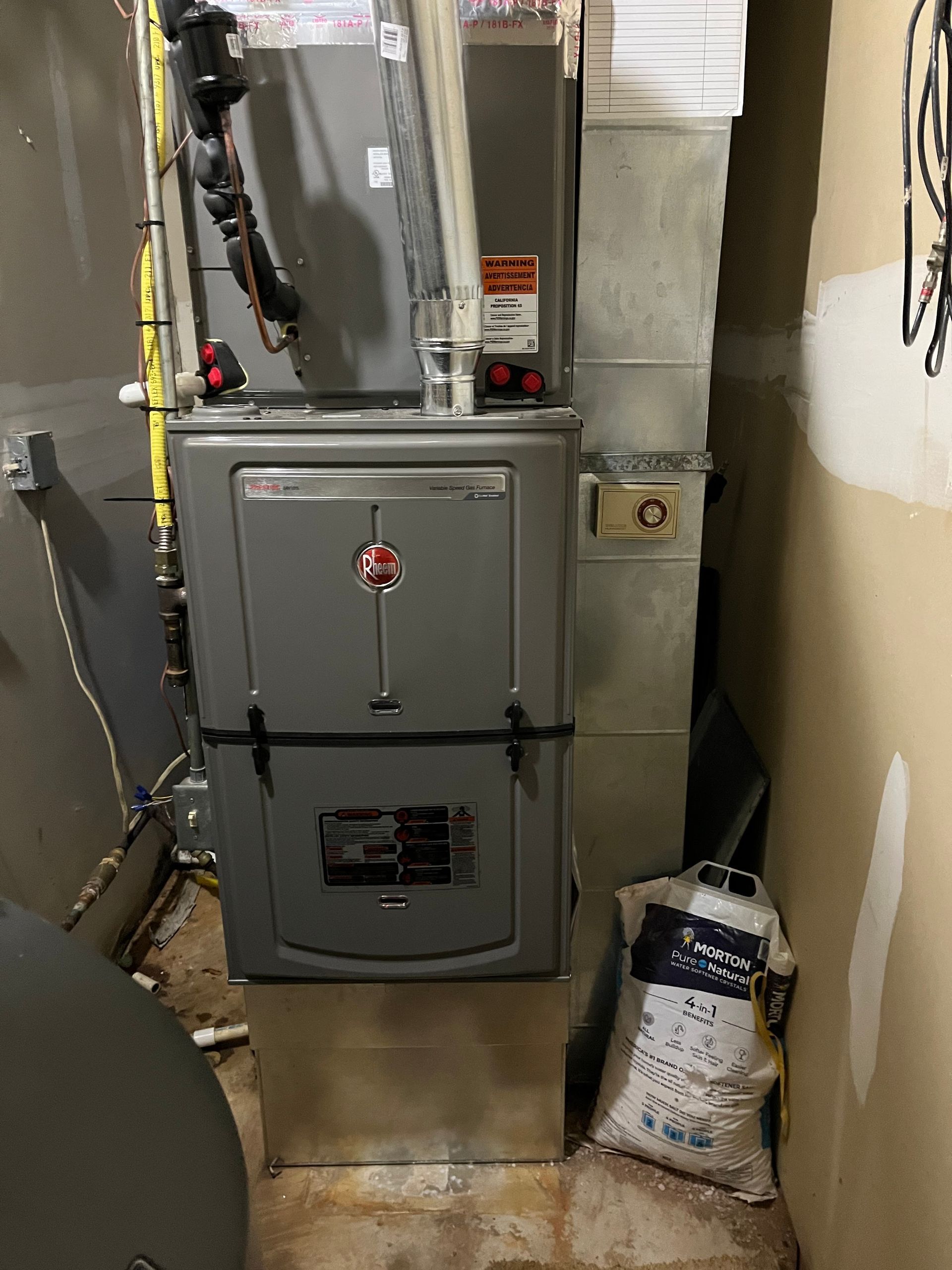 A gray Rheem furnace in a basement setting with ductwork and a bag of material.