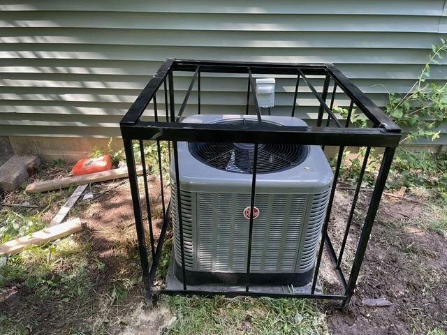 An air conditioning unit encased in a black metal cage, outdoors next to a building with green siding.