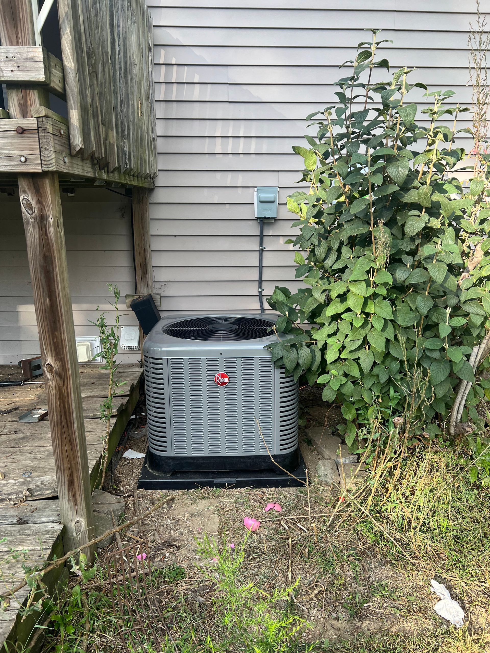 AC unit against a gray-sided building, near a deck and overgrown greenery.