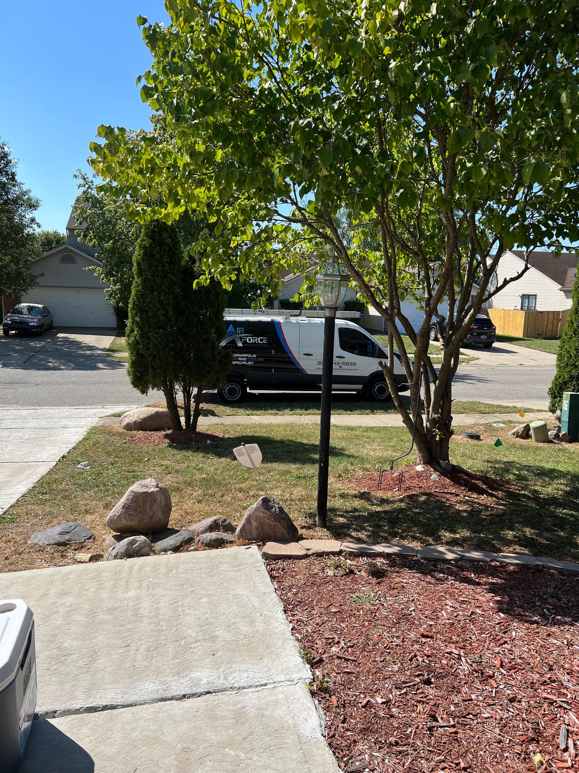 A white and black service van parked on a residential street next to a house with trees and a light post.