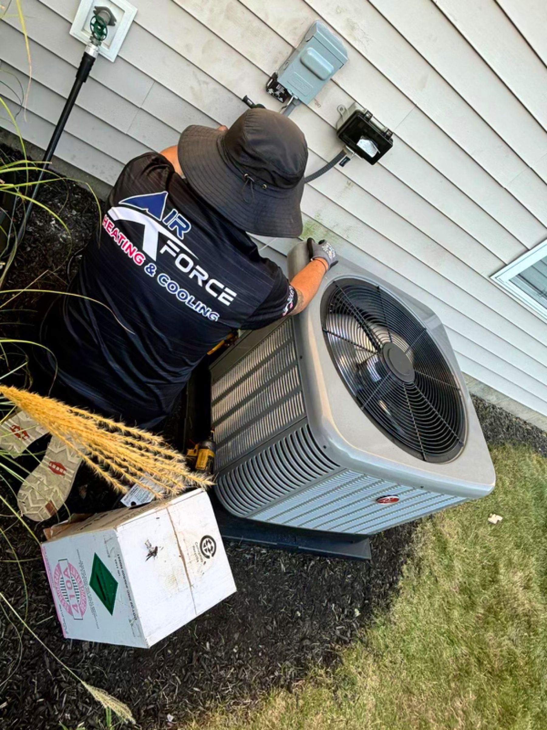 Person in Air Force shirt working on an AC unit near a house, wearing a hat.