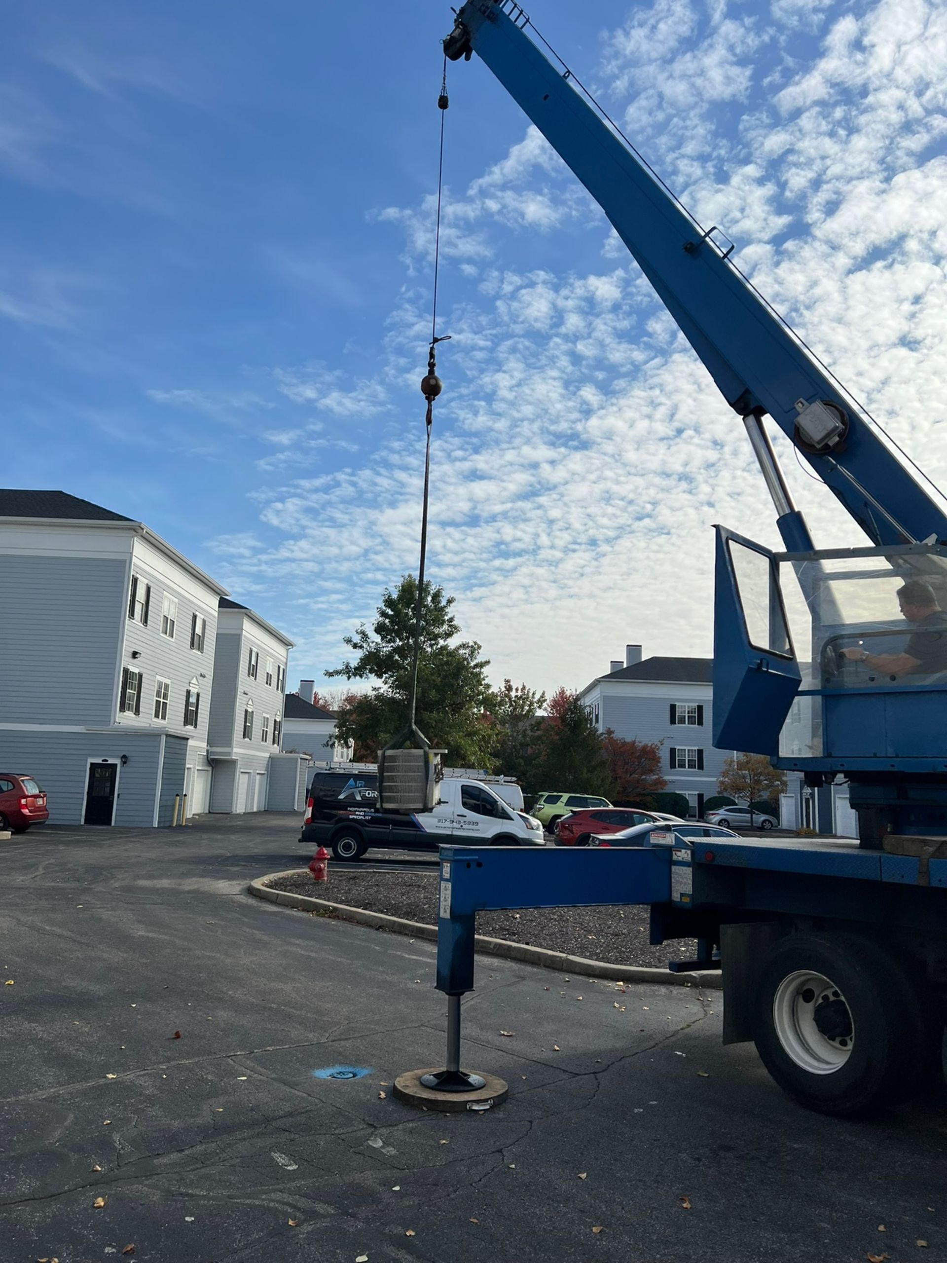 Blue crane lifting something at a parking lot with apartment buildings under a partly cloudy sky.