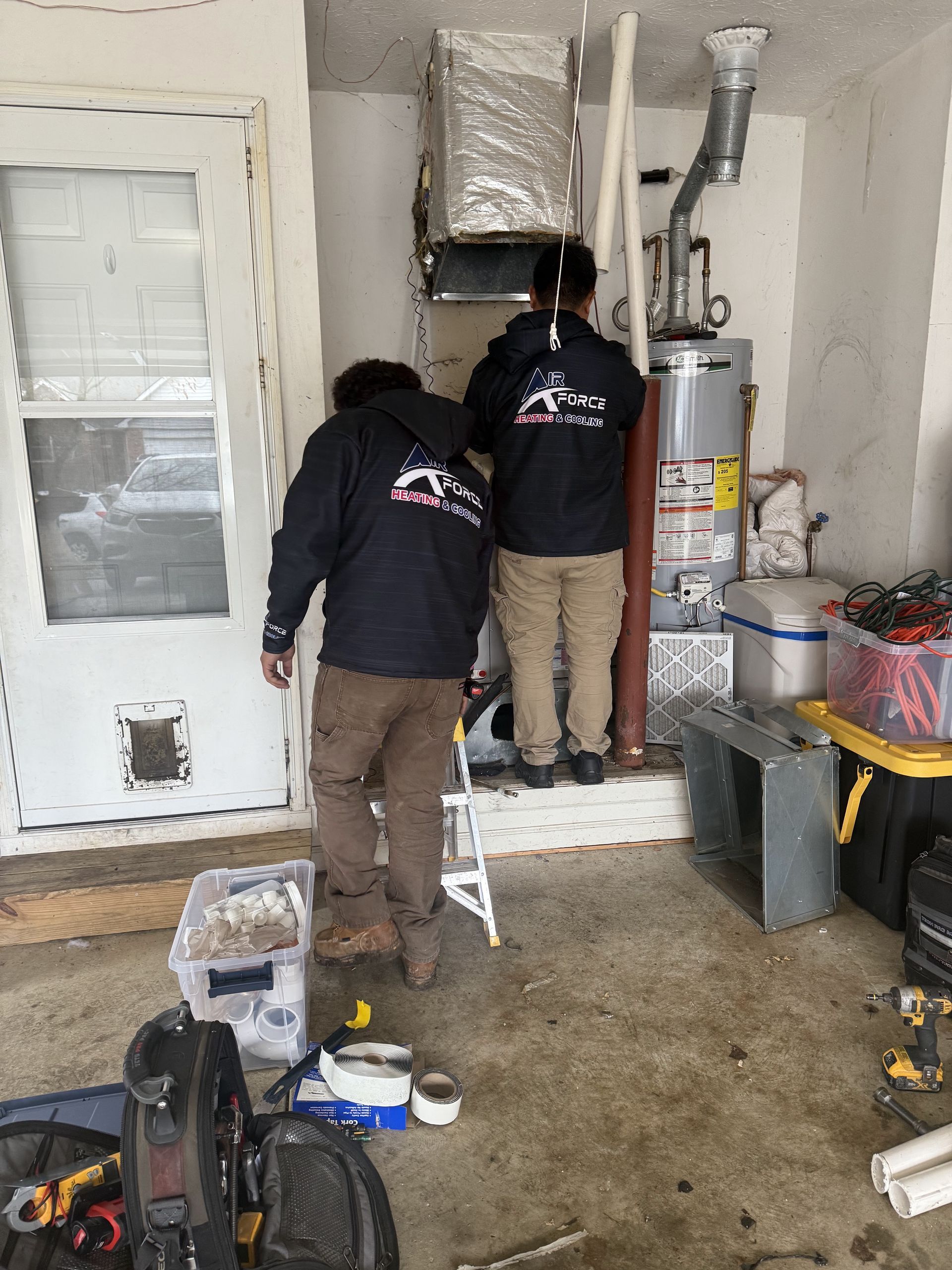 Two technicians in black jackets working on HVAC equipment in a garage.