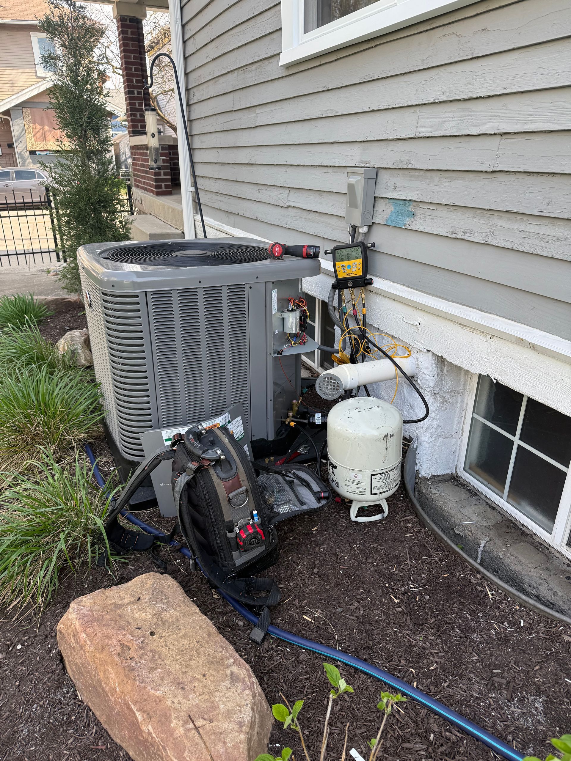 HVAC unit next to a house with a propane tank and a small window in a flowerbed.