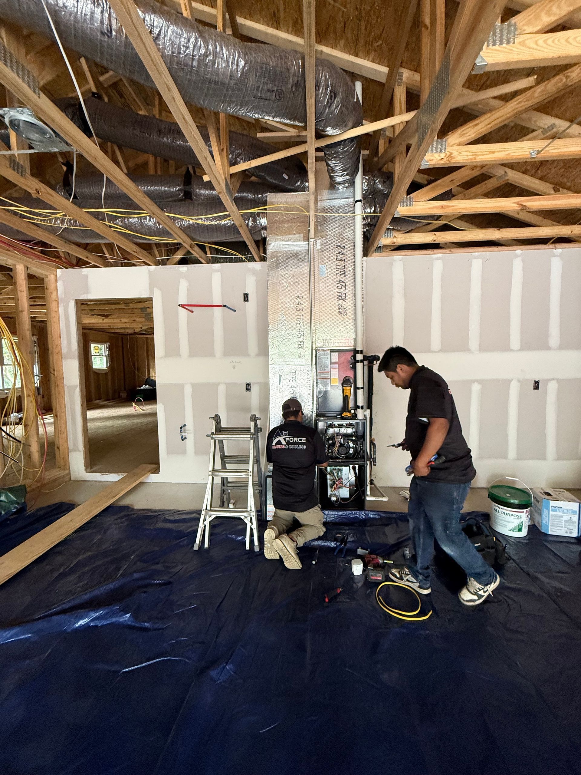 Two workers installing HVAC unit in a room under construction with drywall and exposed wood framing.
