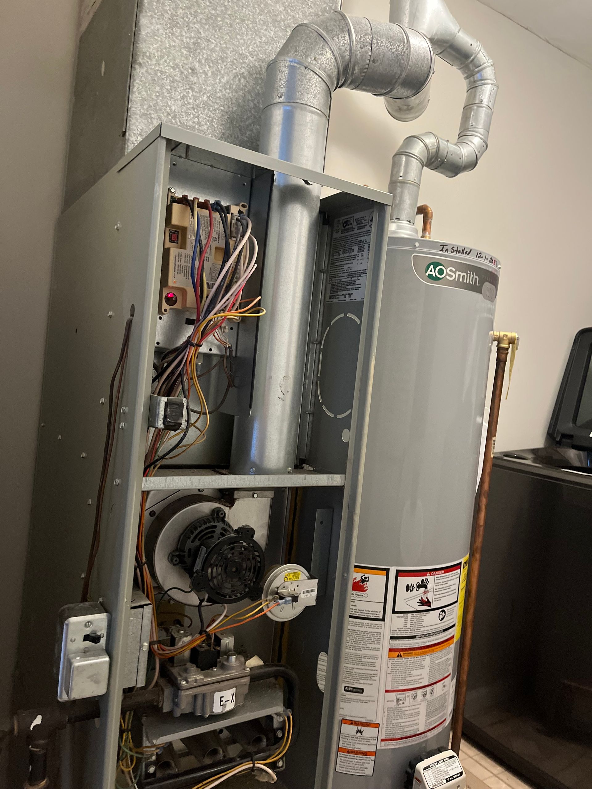 Furnace with open panels next to a hot water heater. Electrical wiring visible. Silver and gray tones.