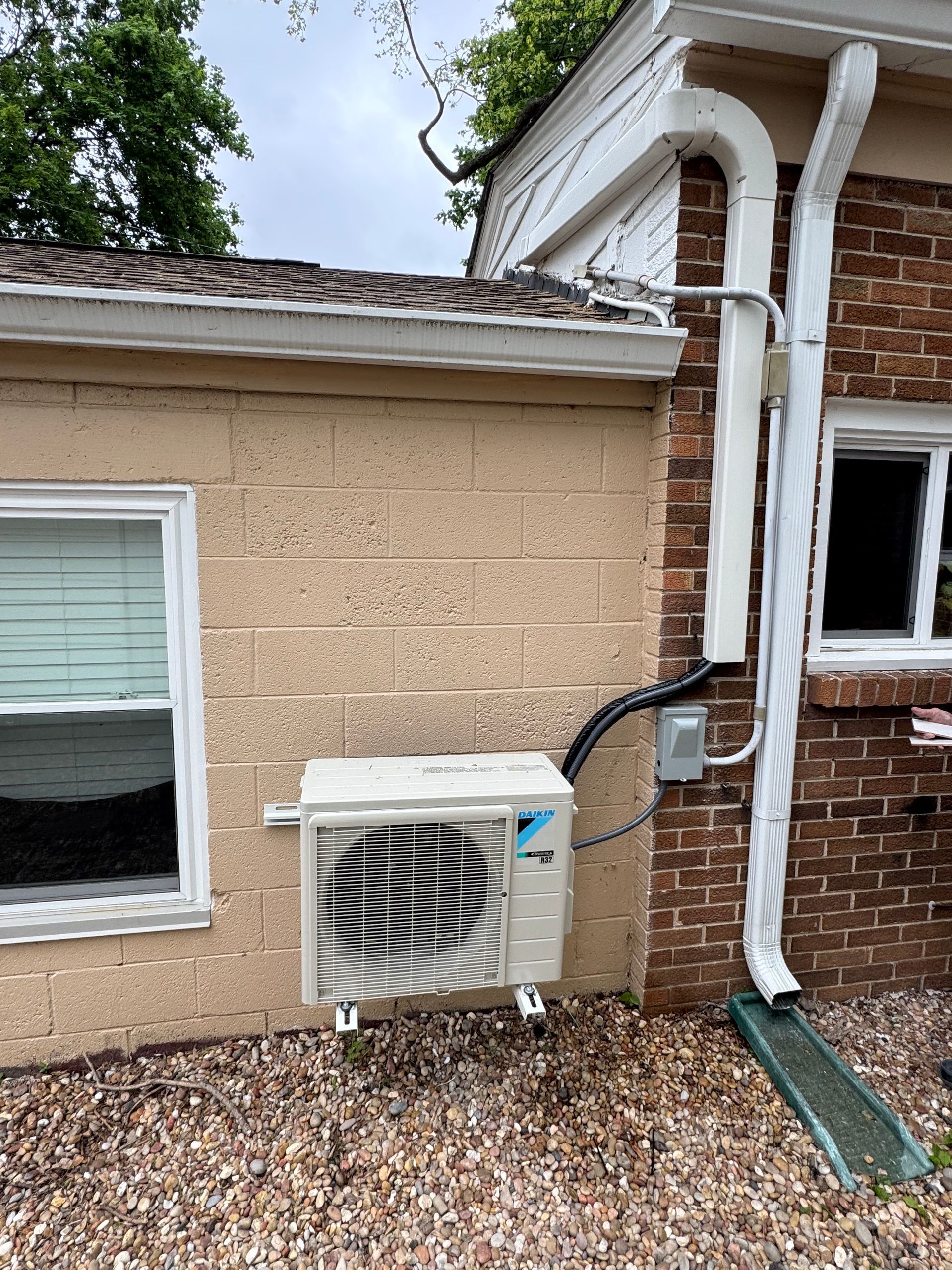 AC unit mounted on a brick wall, next to a window and downspout.