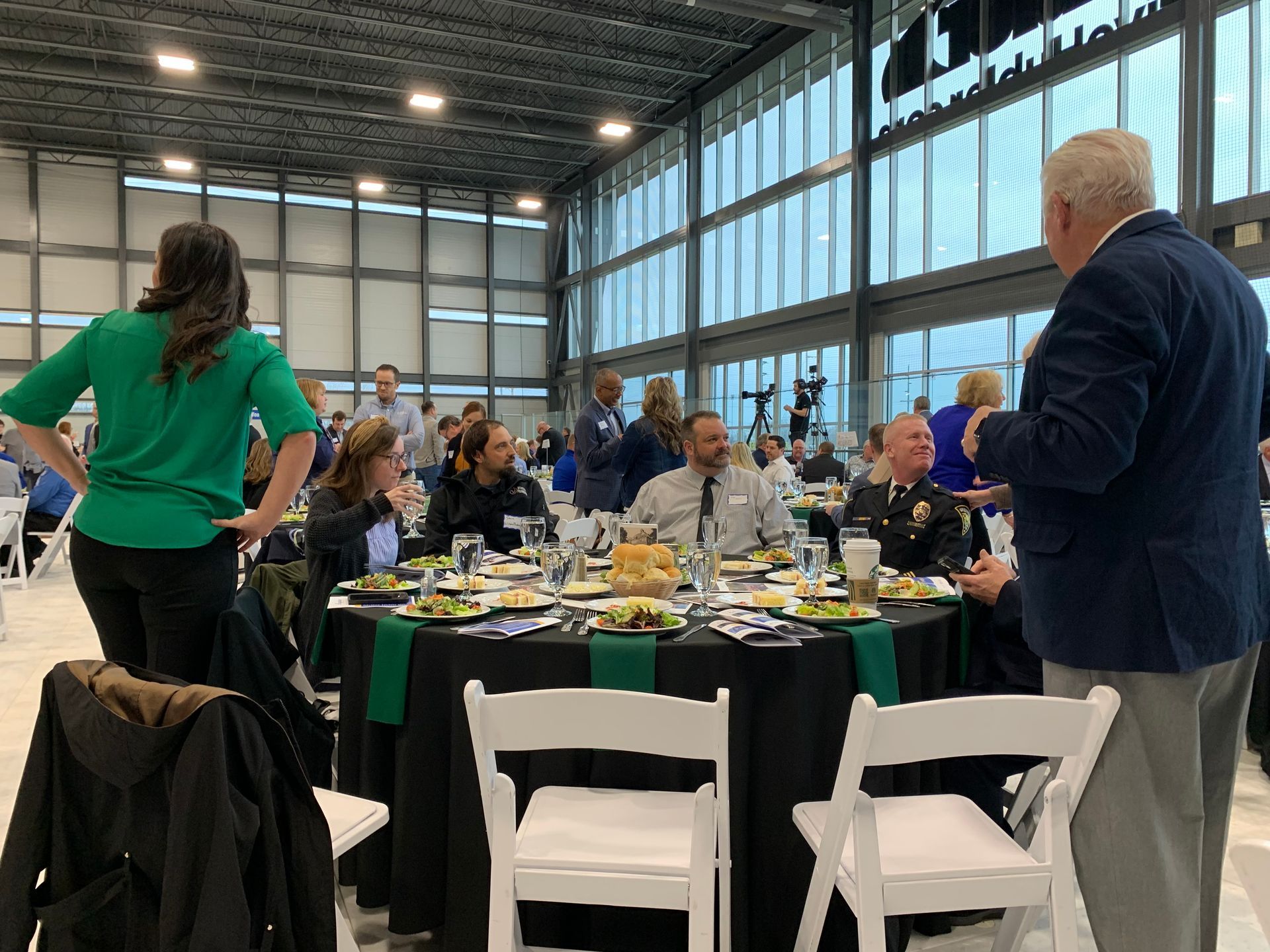 People seated at tables during an event in a modern hall; speaker gesturing.