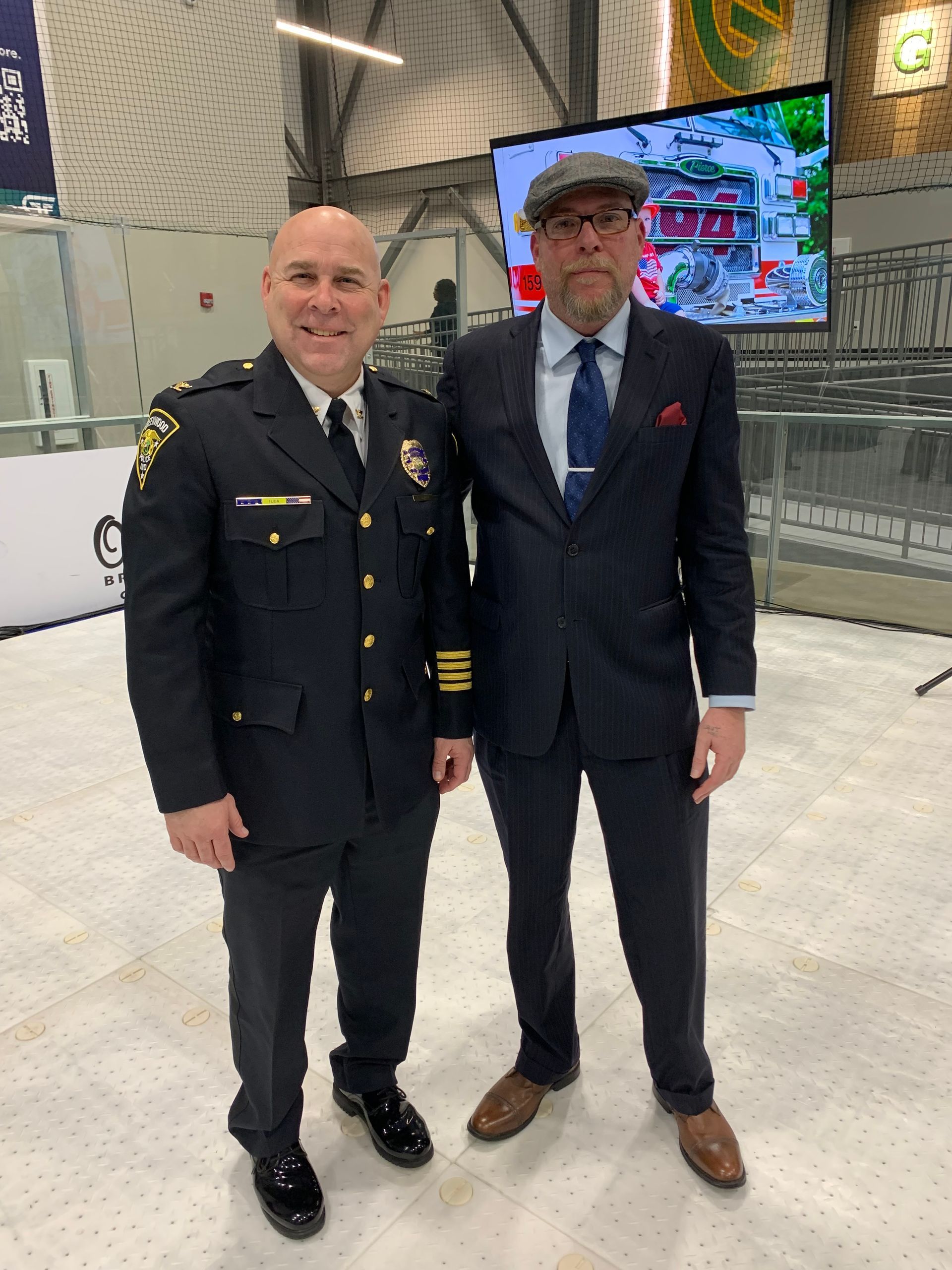 Man in police uniform stands next to a man in a suit, in front of a screen displaying a building.