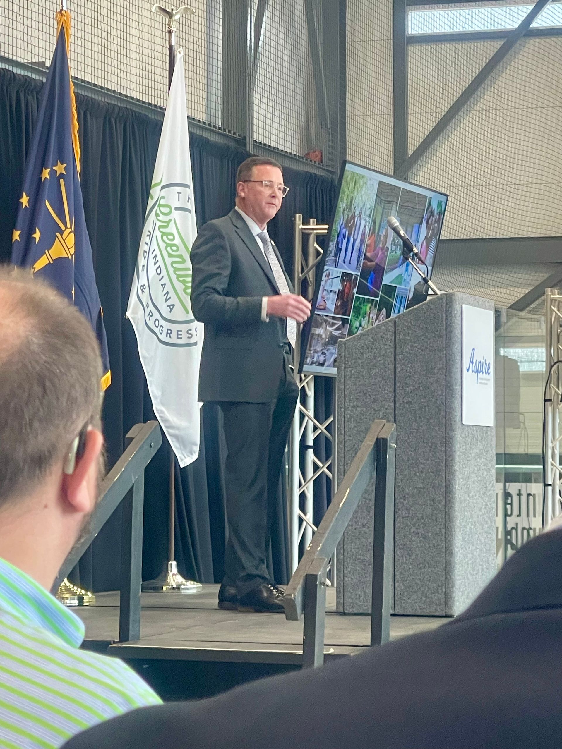 Man in suit speaking at podium, flags behind. Large screen displays photo, indoor setting.