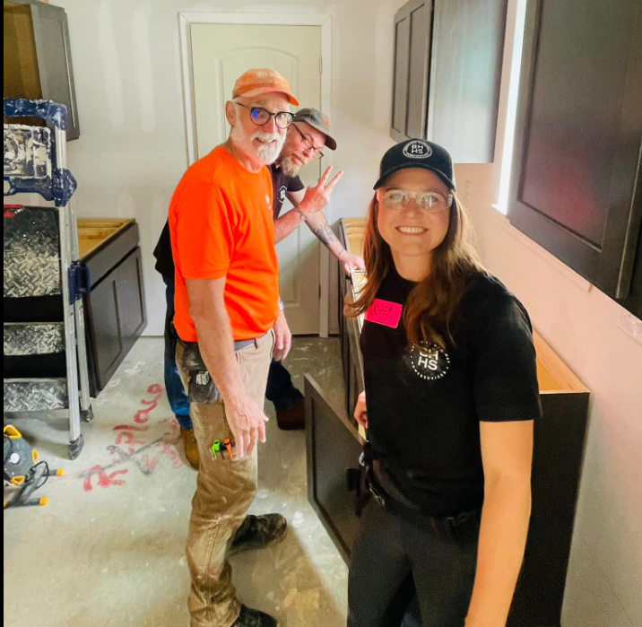 Three people installing kitchen cabinets in a room with tools and unfinished walls.