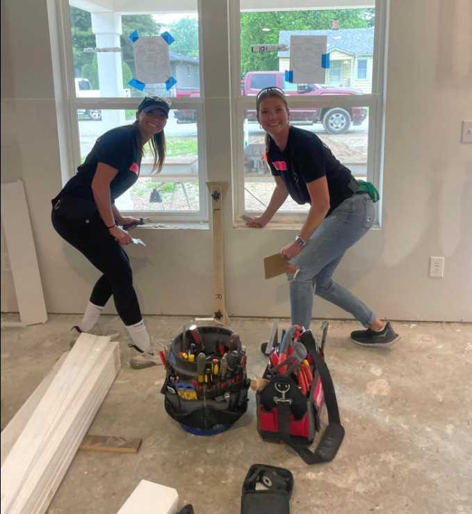 Two women working on window trim in a construction site, tools visible.