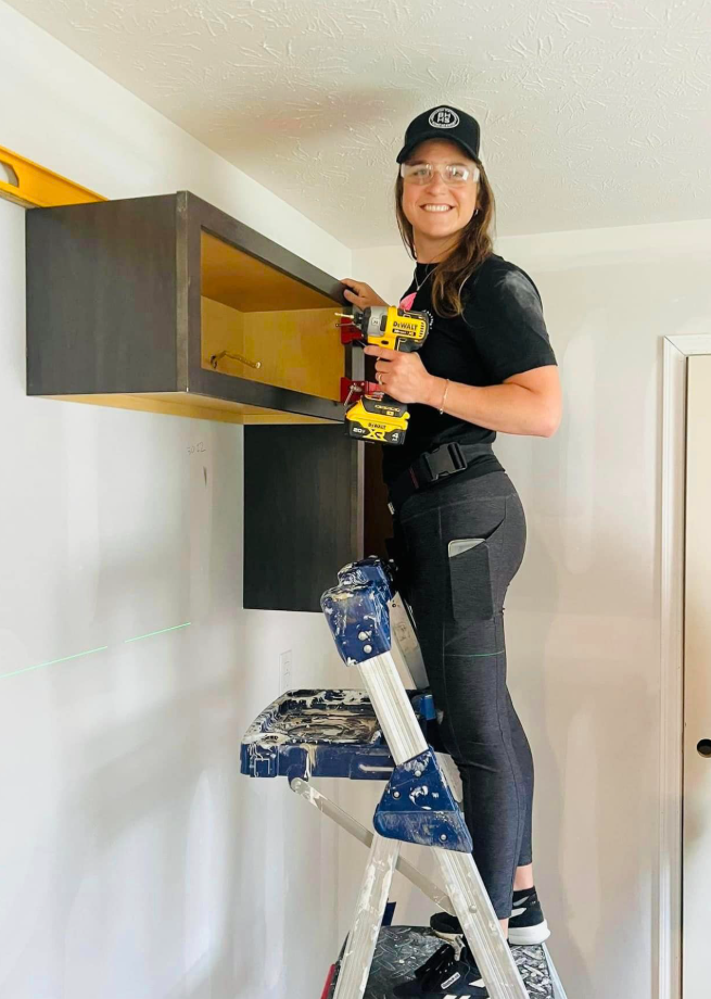 Woman on a ladder installing a dark cabinet with a drill. Bright yellow cabinet interior, level on top.