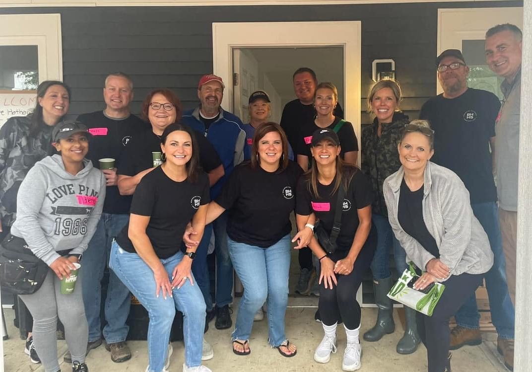 Group of people posing together on a porch, smiling. Many are wearing matching shirts.
