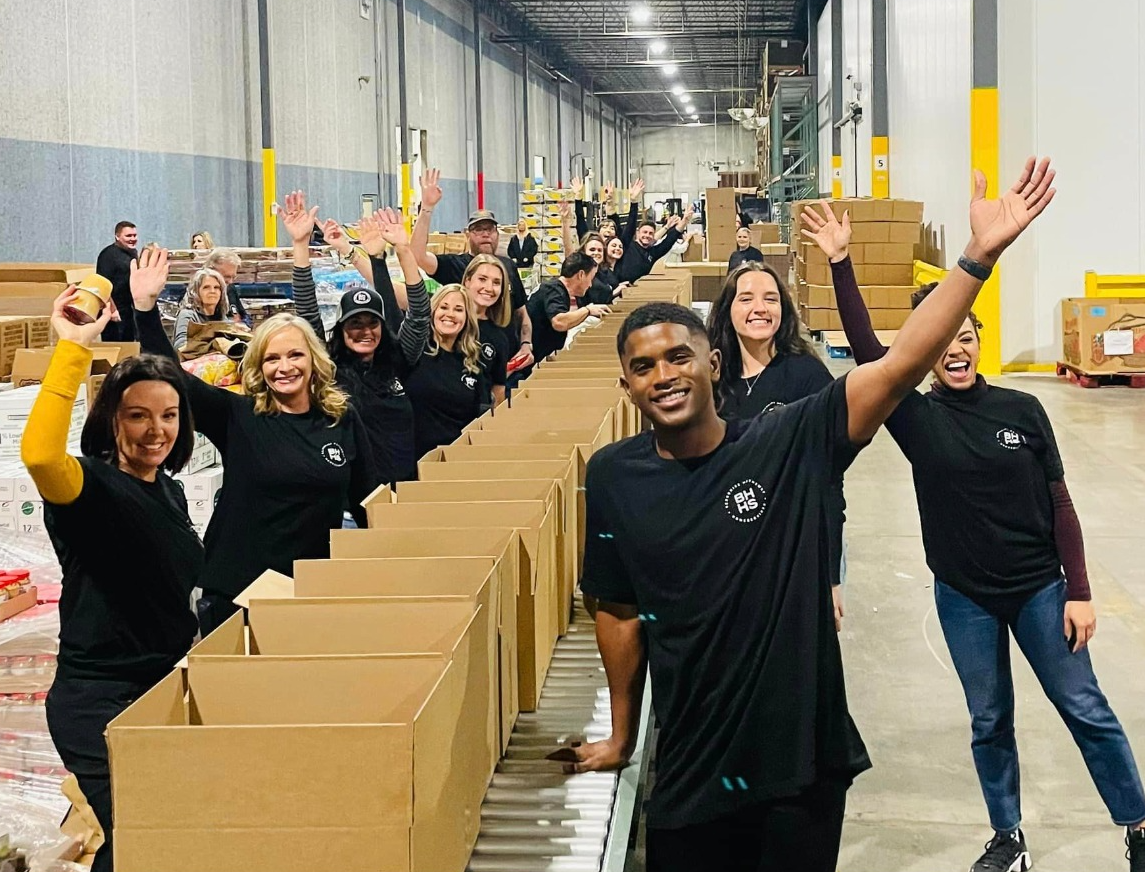 People in black shirts wave next to cardboard boxes in a warehouse.