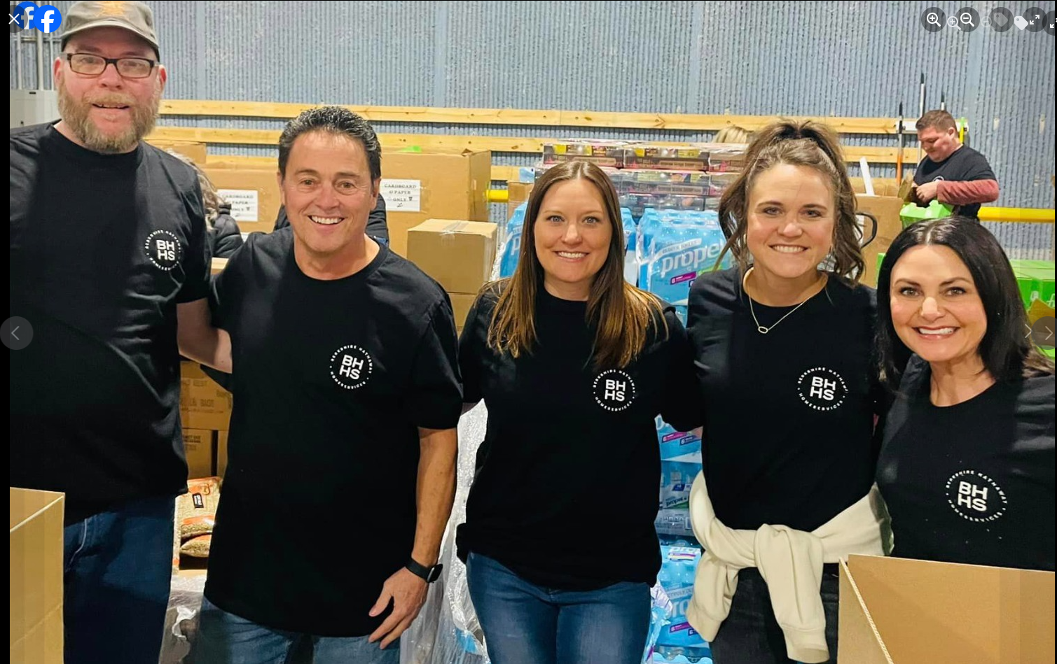 Five people in black t-shirts pose in a warehouse, smiling. Boxes and supplies are in the background.