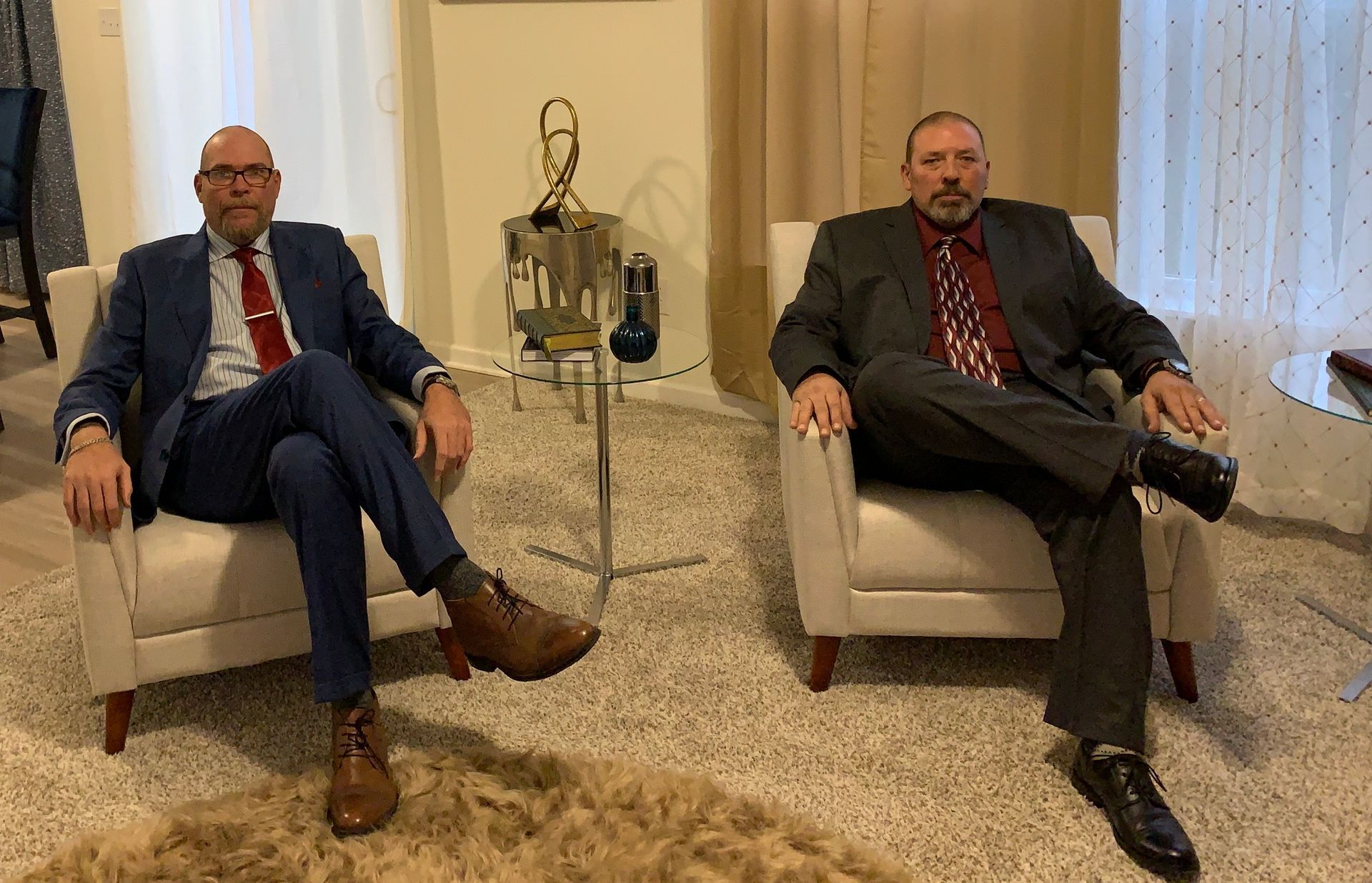 Two men in suits seated in armchairs; indoor setting with neutral tones and decor.