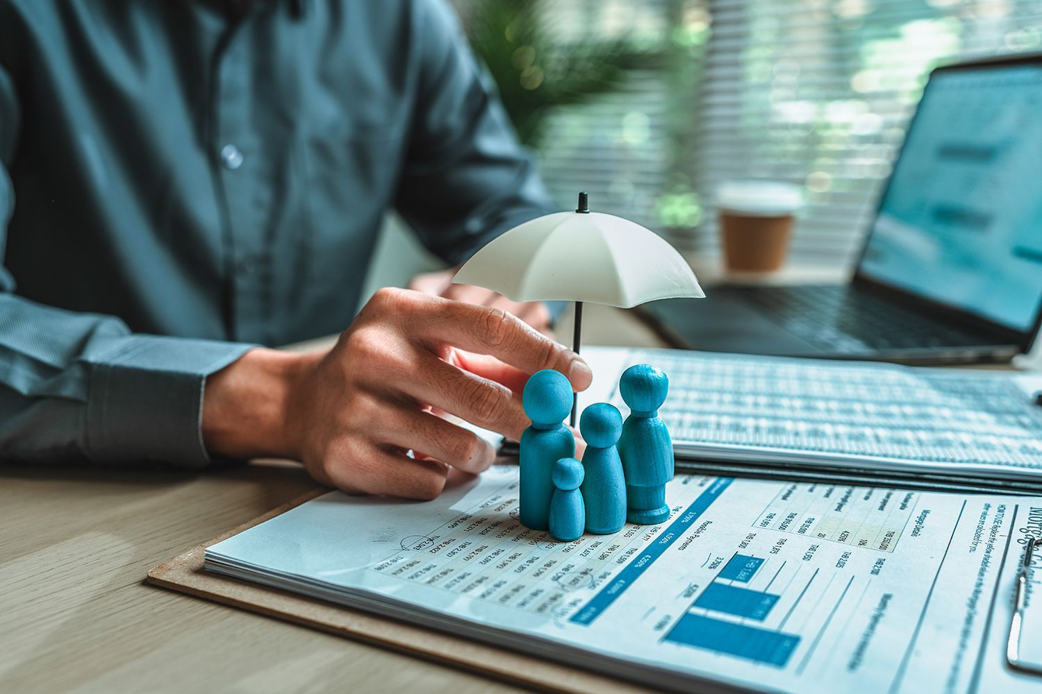 Hand placing miniature figures under a small umbrella on a desk with financial charts and a laptop Hand placing miniature figures under a small umbrella on a desk with financial charts and a laptop