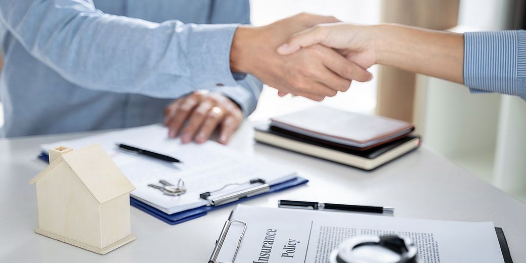 Two people shaking hands over a desk with paperwork, notebooks, and a small house model Two people shaking hands over a desk with paperwork, notebooks, and a small house model