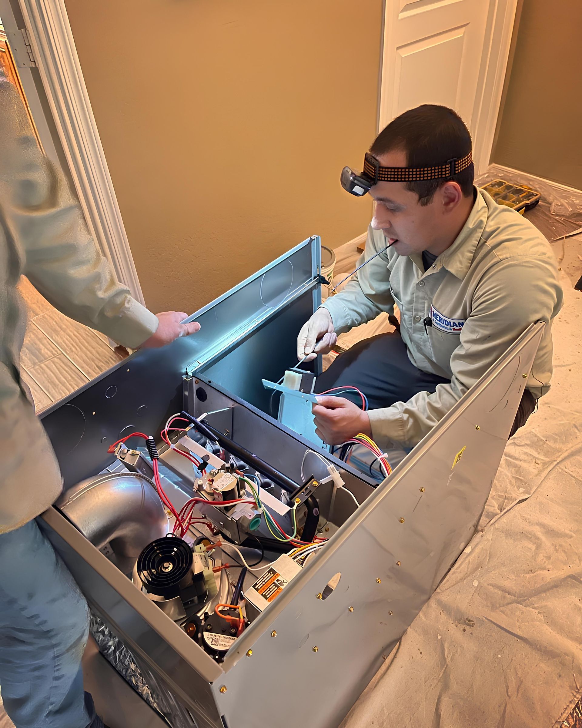 Two technicians working on HVAC unit in a room; one wearing a headlamp, crouched inside and working on wiring.