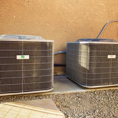 Two air conditioning units side-by-side against a tan wall, on a concrete slab, with gravel ground.