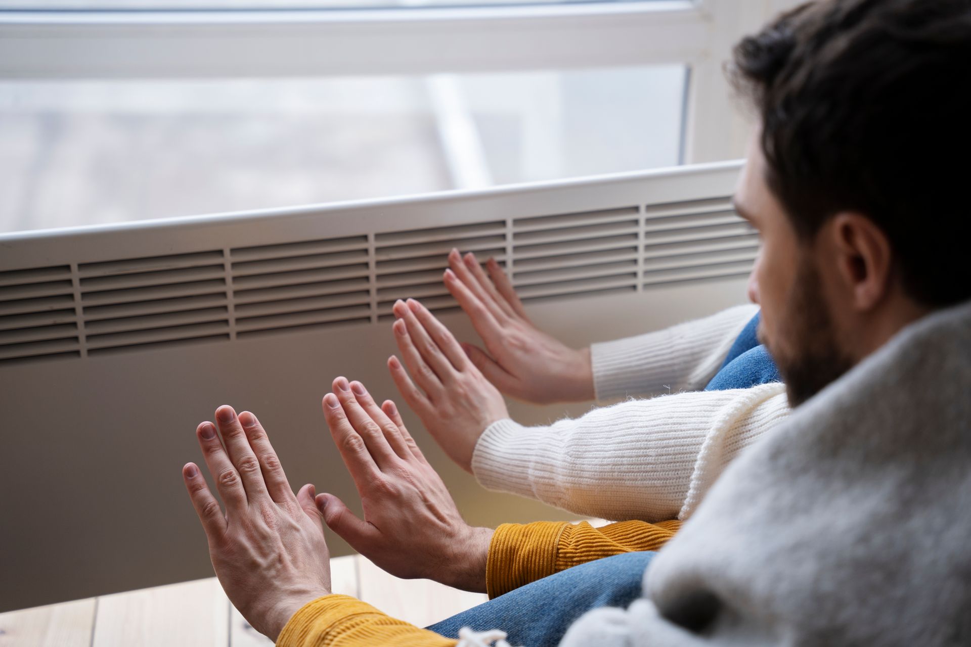 A man and a woman are warming their hands in front of a radiator.
