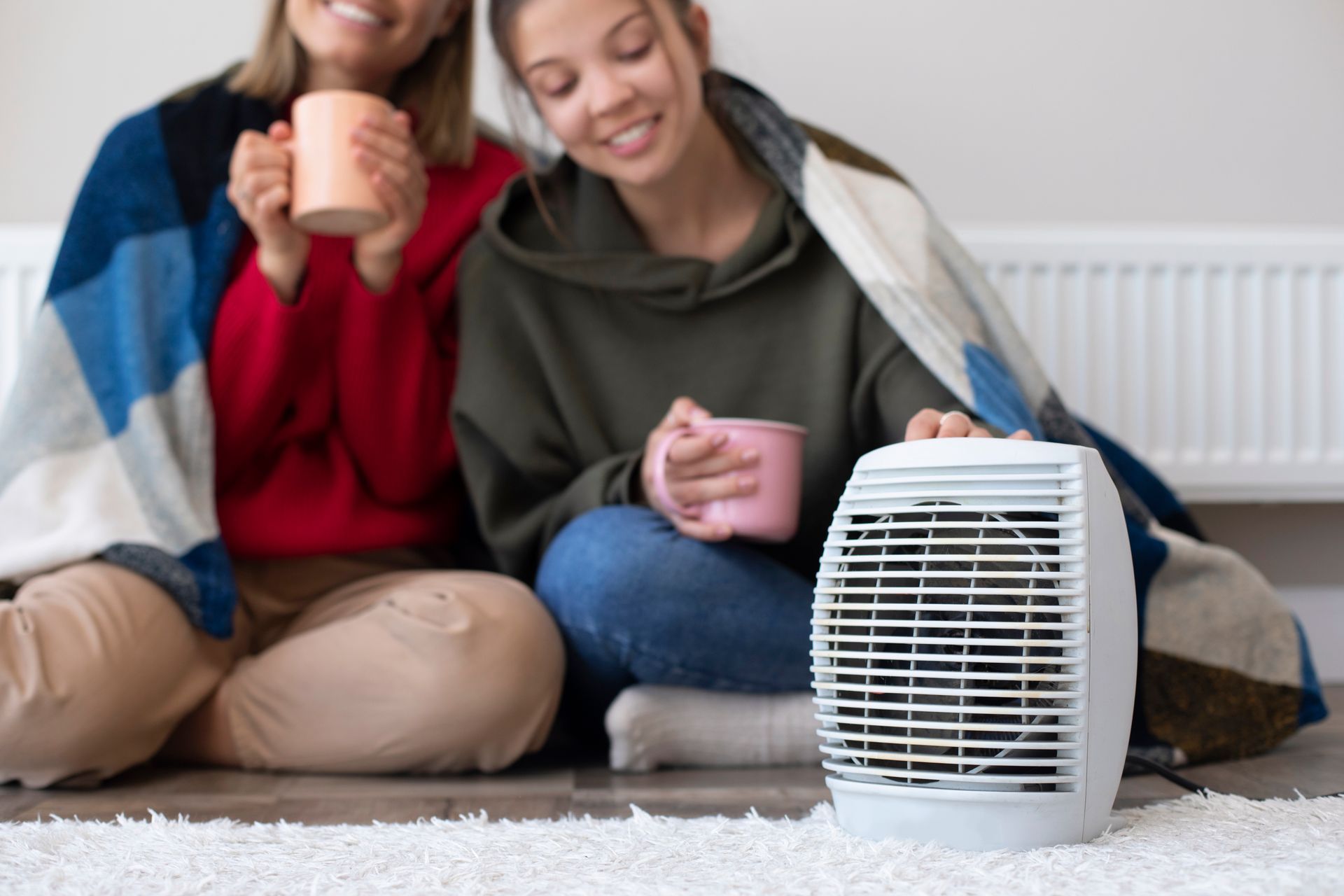 Two women are sitting on the floor with cups of coffee and a heater.