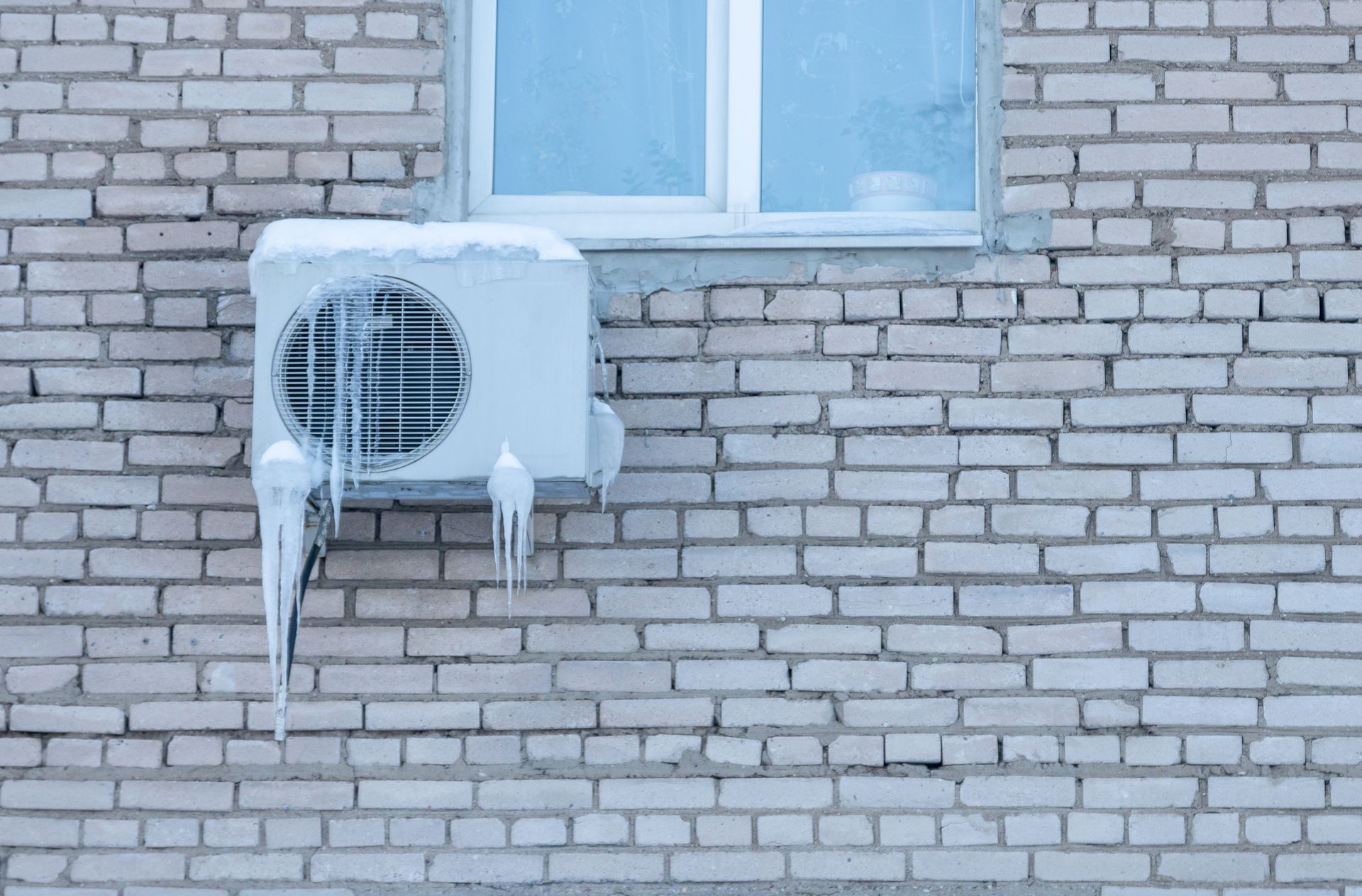 Icy outdoor air conditioner unit mounted on a brick wall beneath a window