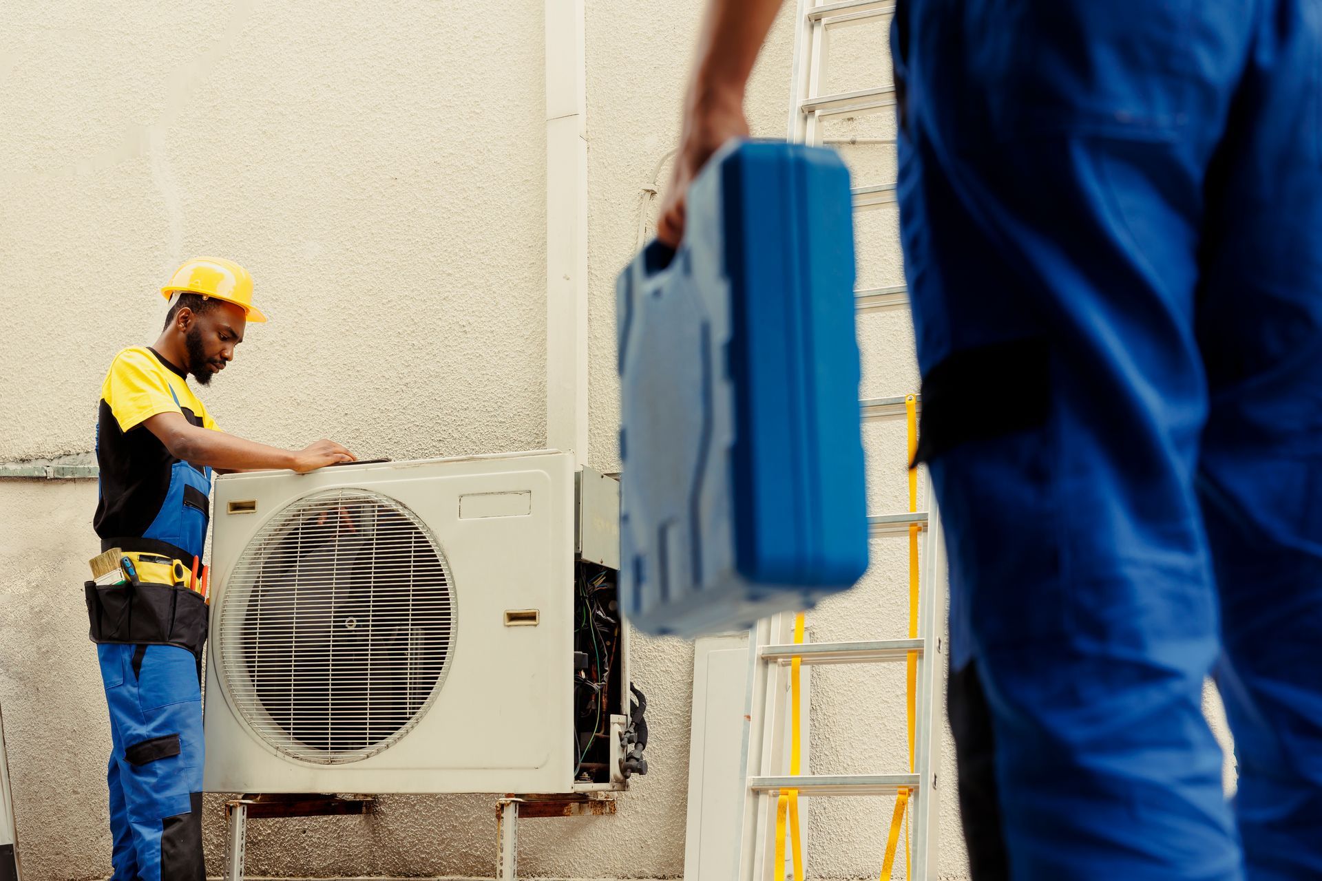Two HVAC technicians servicing an air conditioning unit outside. One works on the unit; the other holds a toolbox.