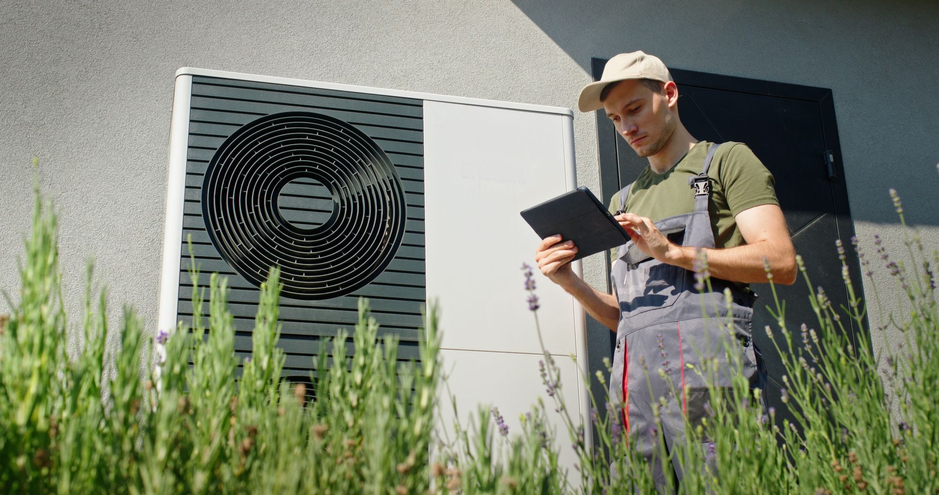 Technician in overalls uses tablet to inspect heat pump outside a house.