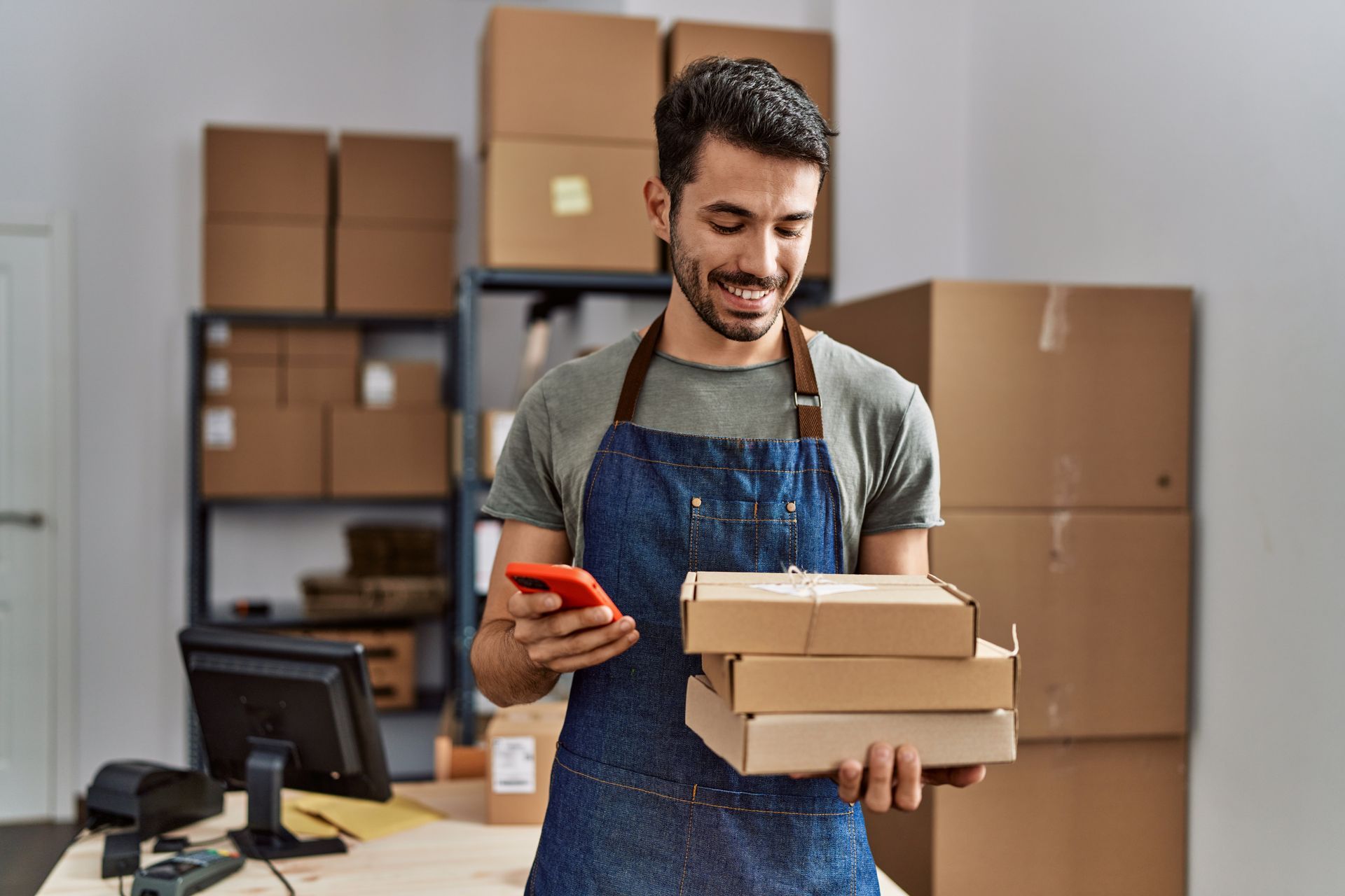 Man in apron smiles, looking at phone, holding stacked packages in a warehouse with boxes. Man in apron smiles, looking at phone, holding stacked packages in a warehouse with boxes.